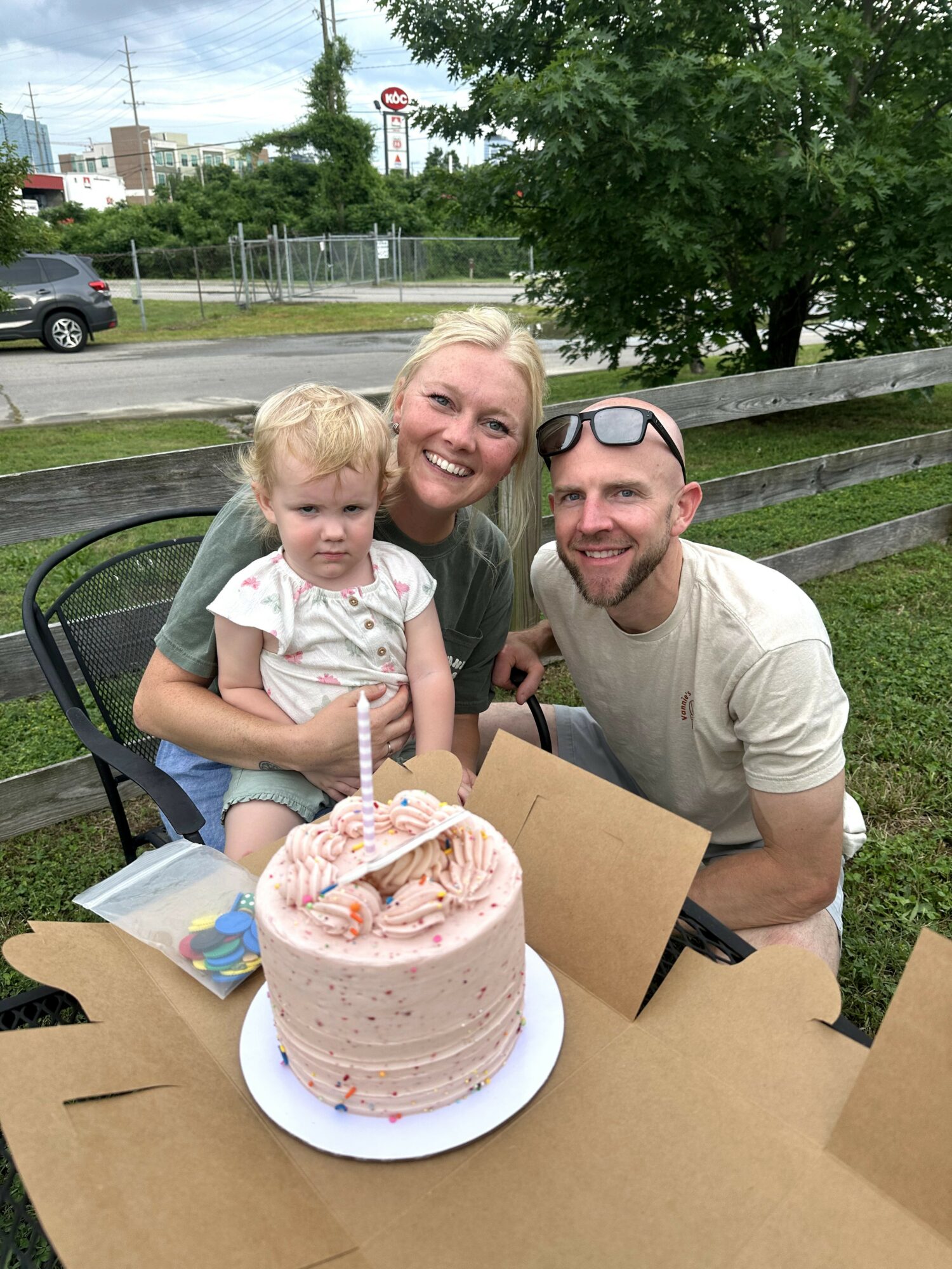 Three people smiling outdoors with a birthday cake, a young girl, a woman, and a man, near a wooden fence.