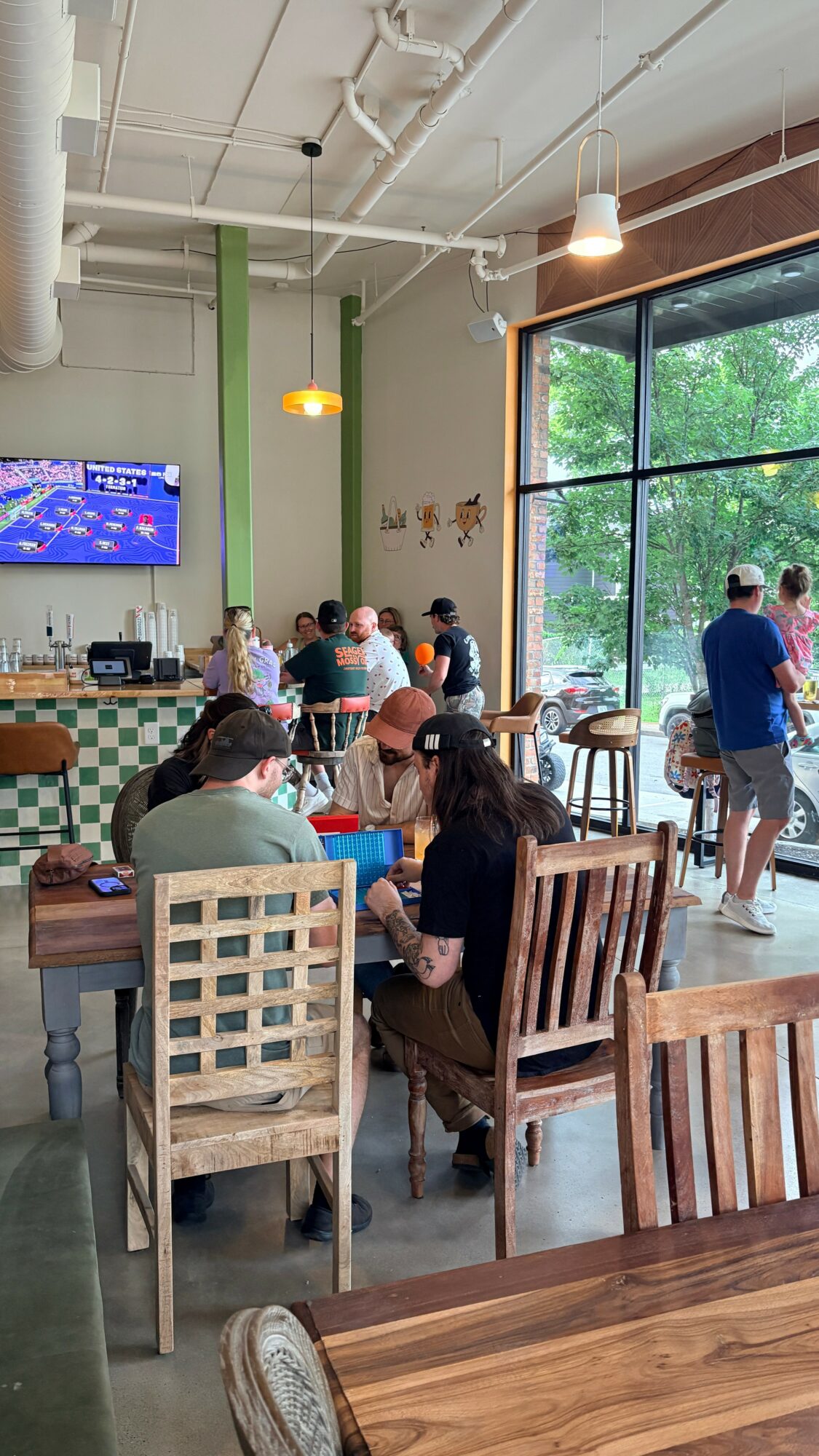 People sit at tables inside a cafe with large windows showing trees outside, some standing near the window.