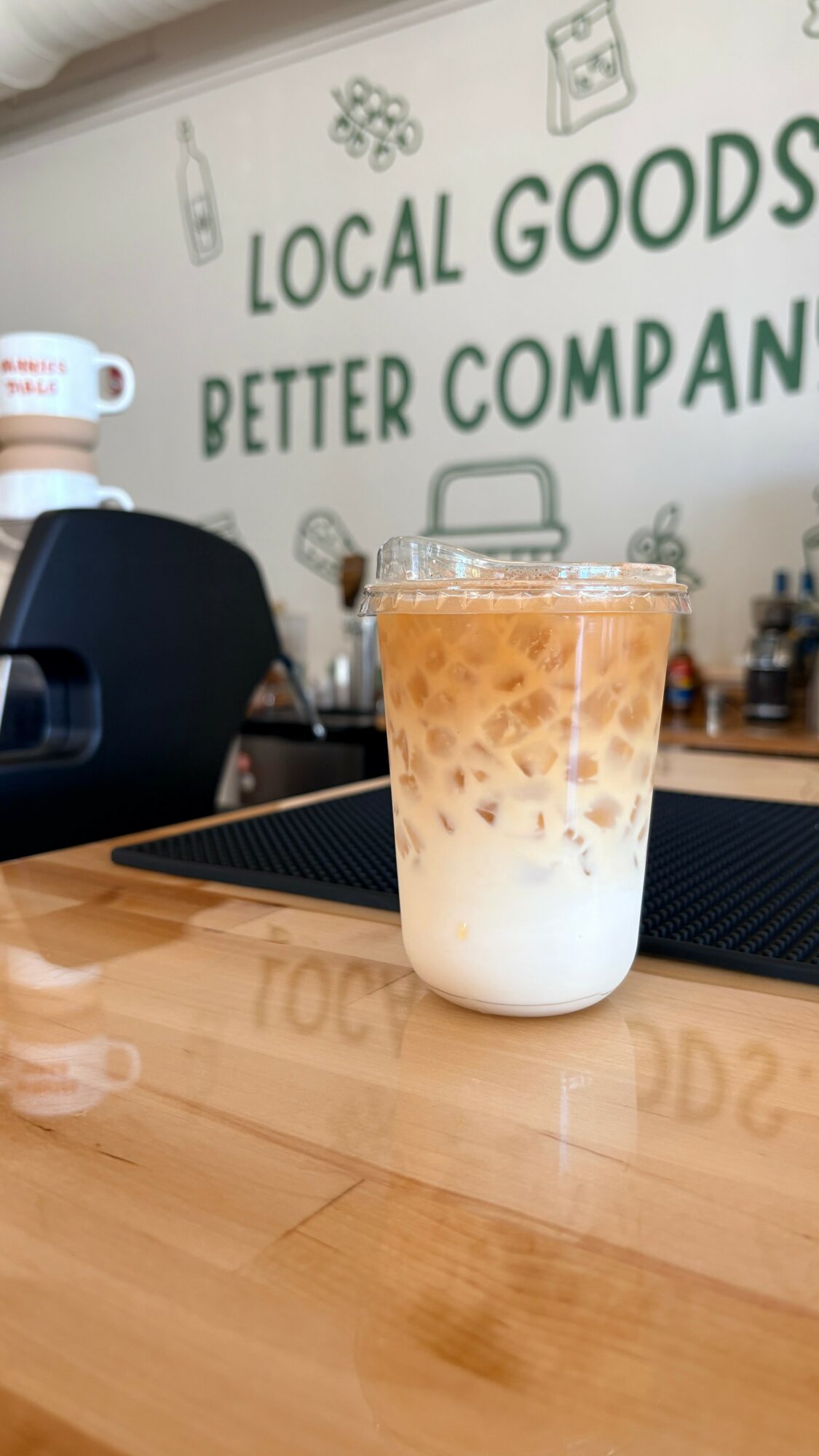 Iced coffee with milk in a clear glass on a wooden table, background shows a sign with text and illustrations.