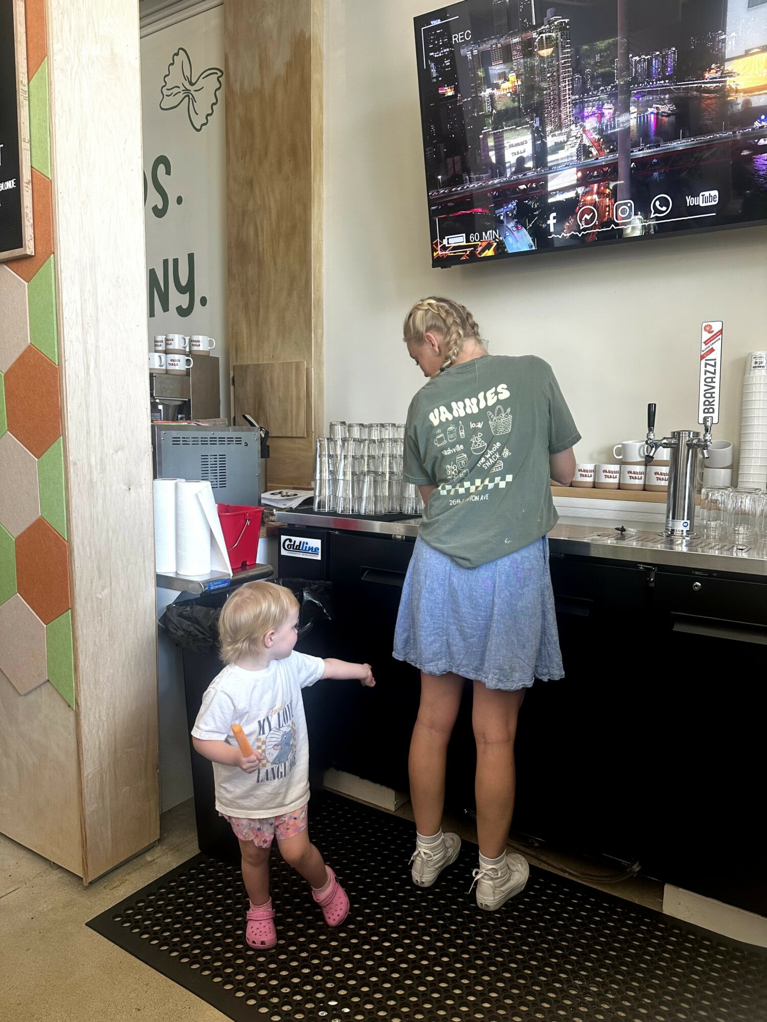 A woman and a young girl stand at a beverage counter with a large TV screen above. The woman is pouring a drink, the girl points at something. The girl wears a white shirt and pink shoes, the woman wears a green shirt and a blue skirt. The setting appears to be a cafe or restaurant.