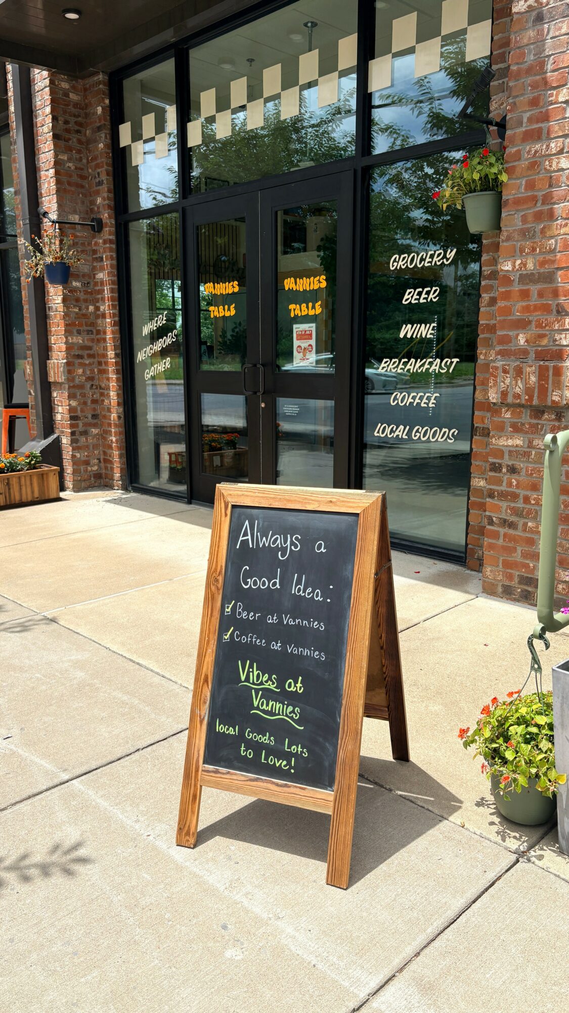 Chalkboard sign outside a cafe with handwritten message, surrounded by potted plants and brick building, sunny day.