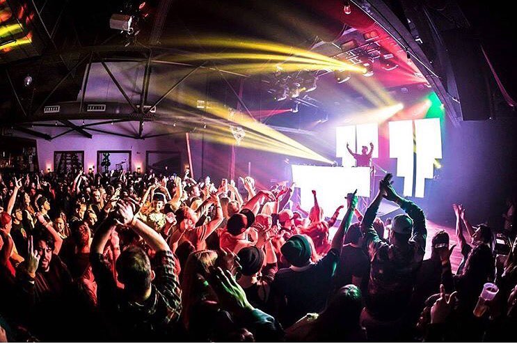 Crowd dancing and raising hands at a concert with colorful stage lights and a DJ on stage.