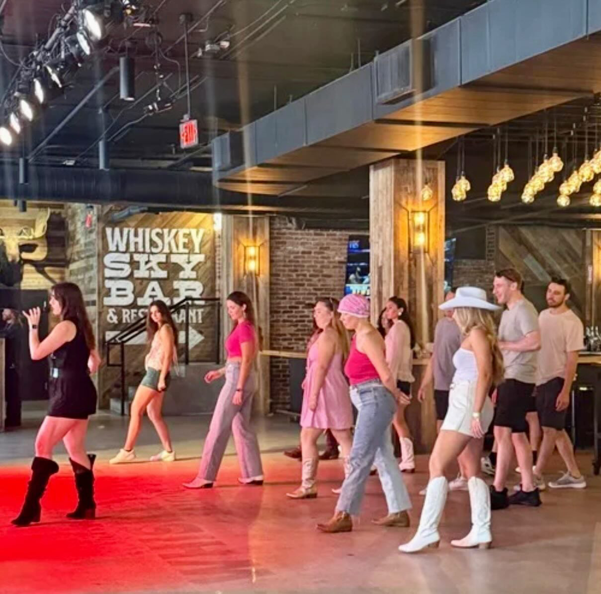 Group of people line dancing in a bar with brick walls and warm lighting, some wearing cowboy hats and boots.
