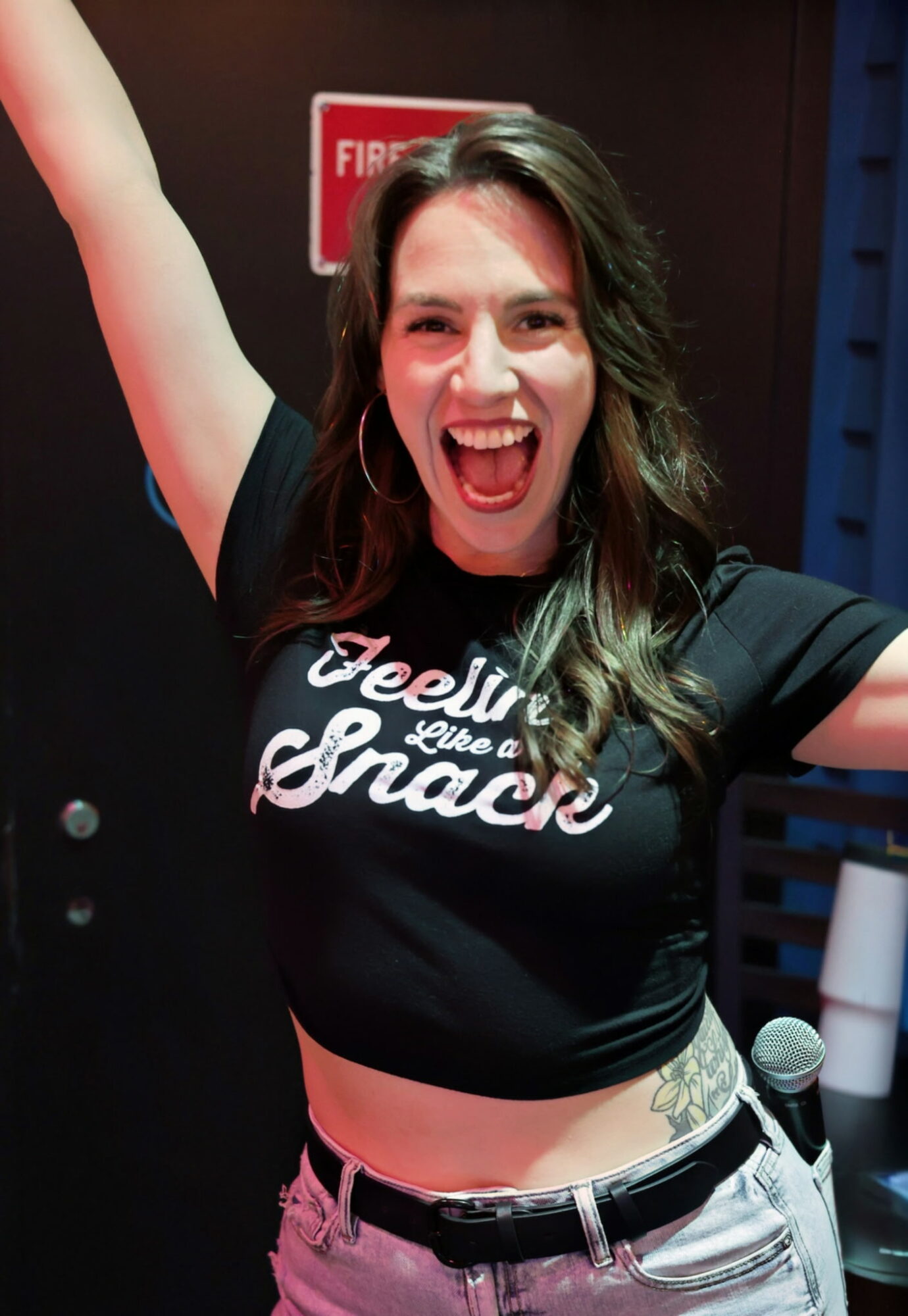Woman with long hair smiling and raising her arm, wearing a black shirt and light-colored pants, in an indoor setting.