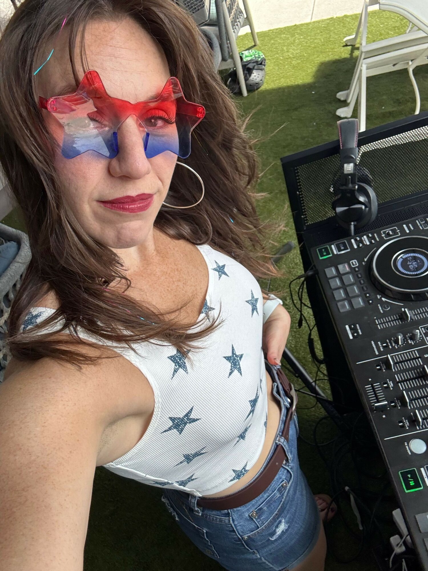 Woman with red glasses and star-patterned shirt standing outdoors near DJ equipment.