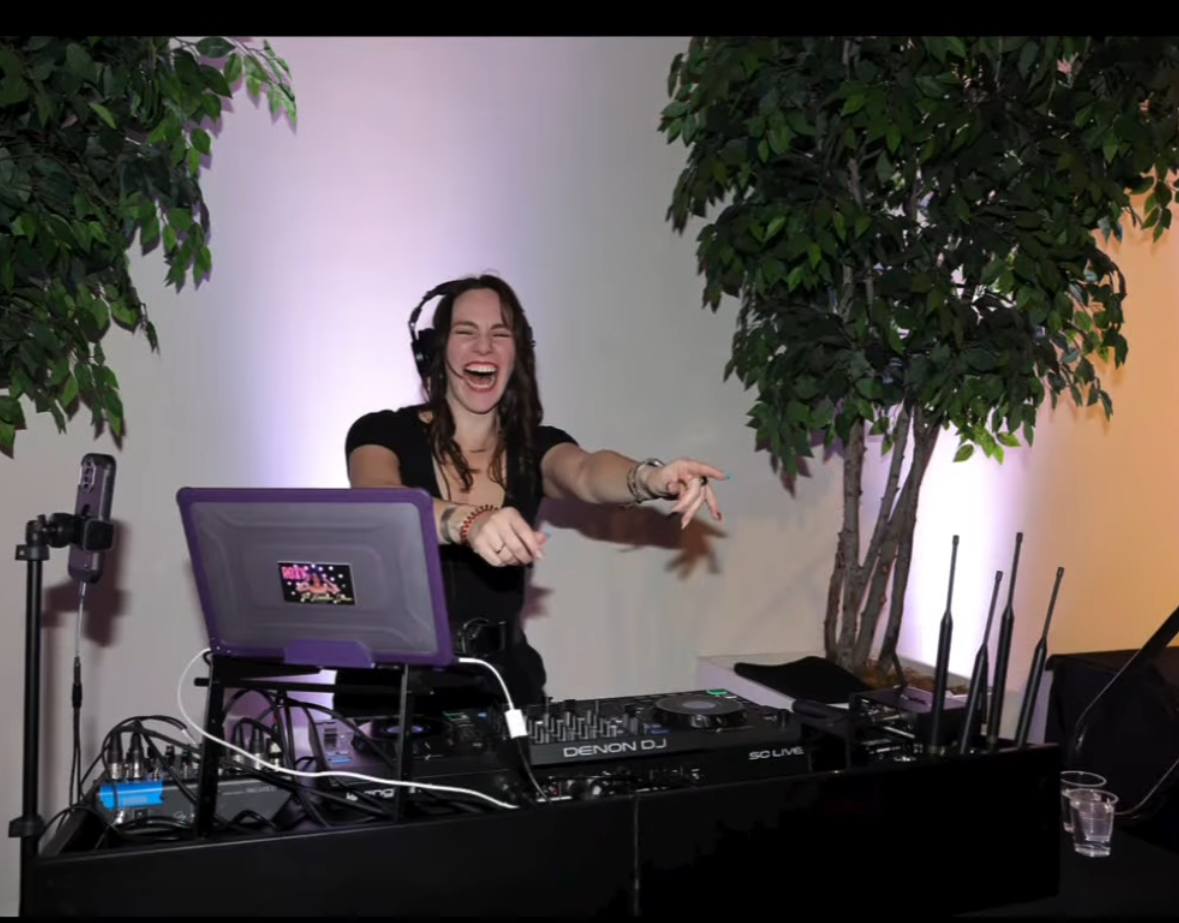 Female DJ smiling and dancing behind a DJ setup with equipment and a laptop, flanked by two large potted plants.