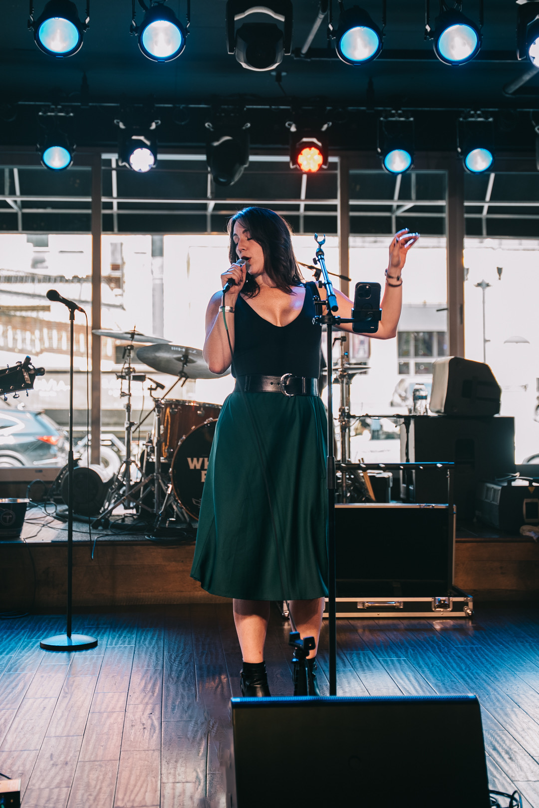 A woman singing on stage with a microphone, wearing a black top and green skirt, in a venue with stage lights.