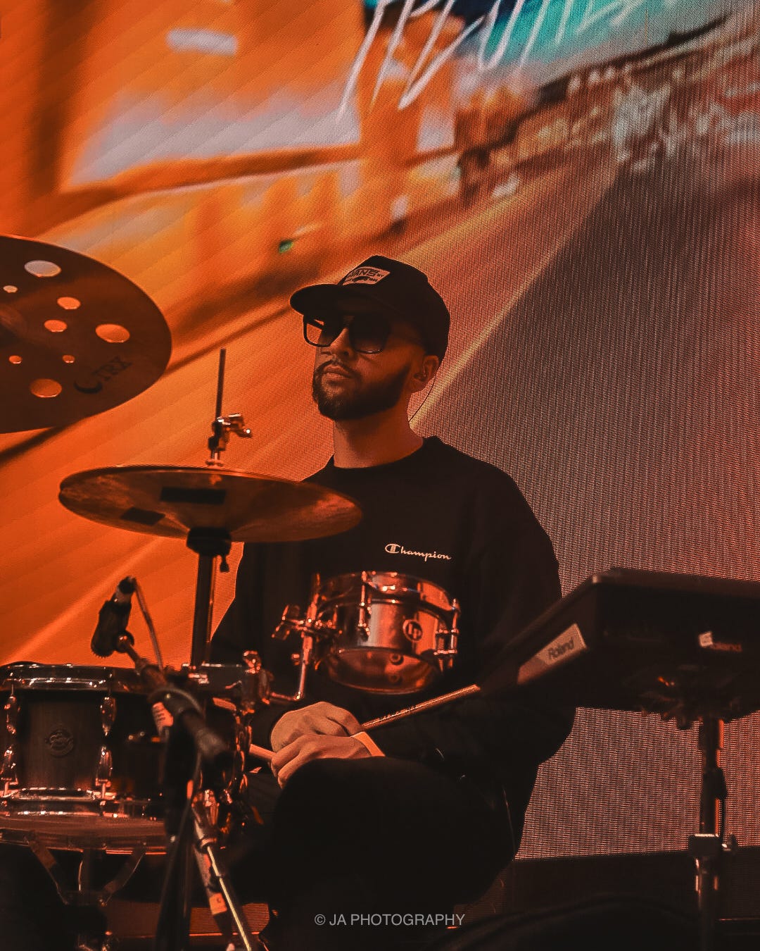 Musician playing drums on stage with colorful background, wearing sunglasses and a cap, focused on performance.