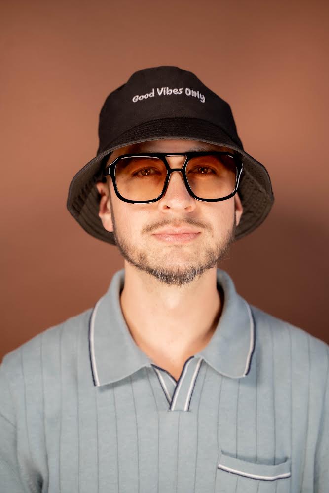 Young man wearing glasses, a bucket hat with 'Good Vibes Only' text, and a light gray polo shirt, smiling slightly.