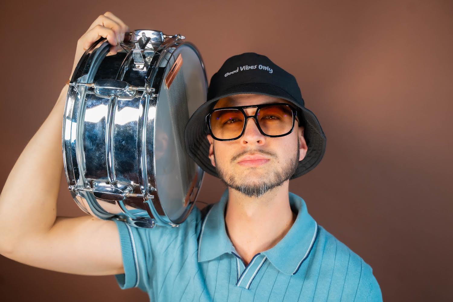 Man wearing sunglasses, bucket hat, and blue polo shirt holding a drum over shoulder, neutral expression, plain background.
