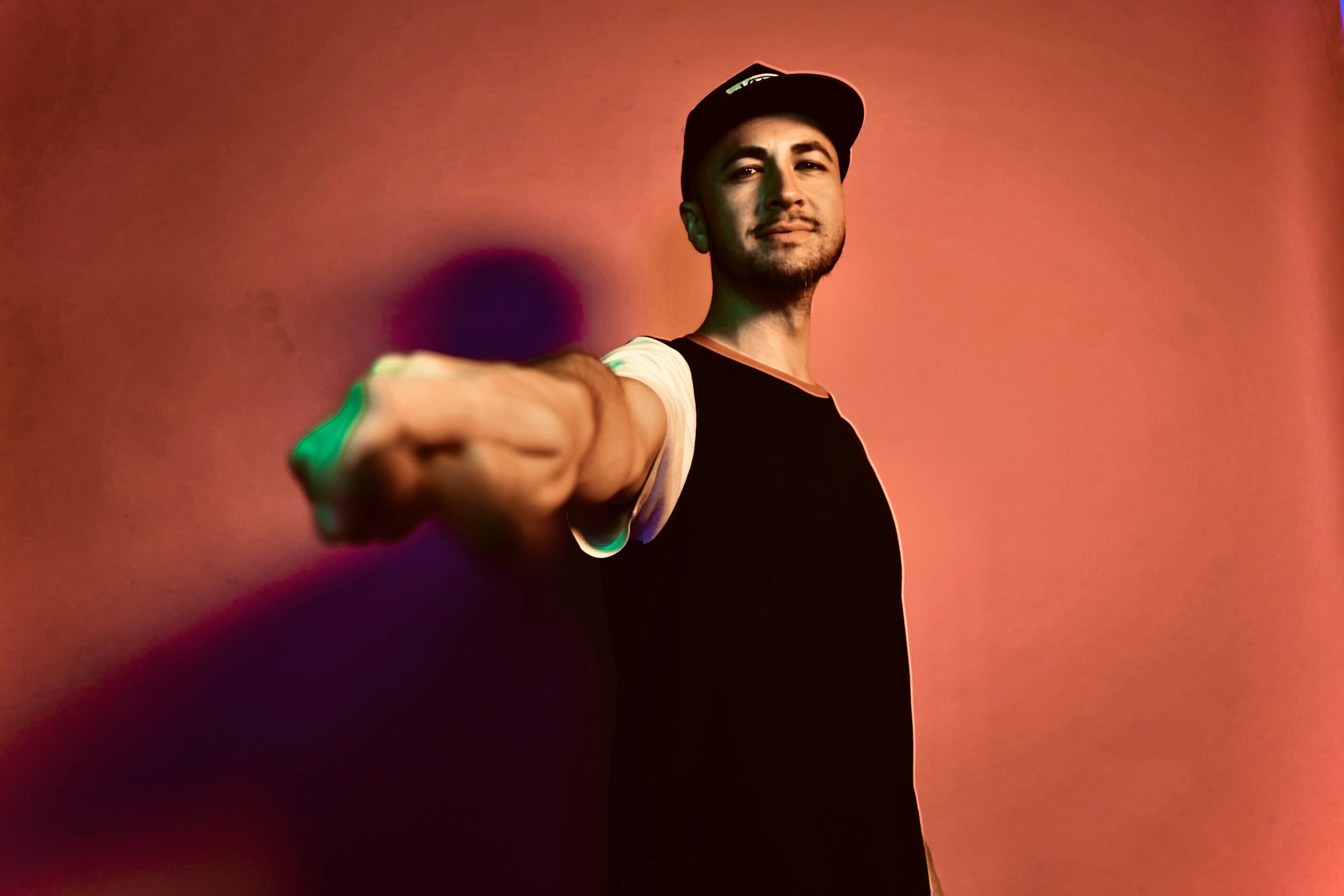 Young man in black shirt and cap extending his arm towards the camera against a pink background.