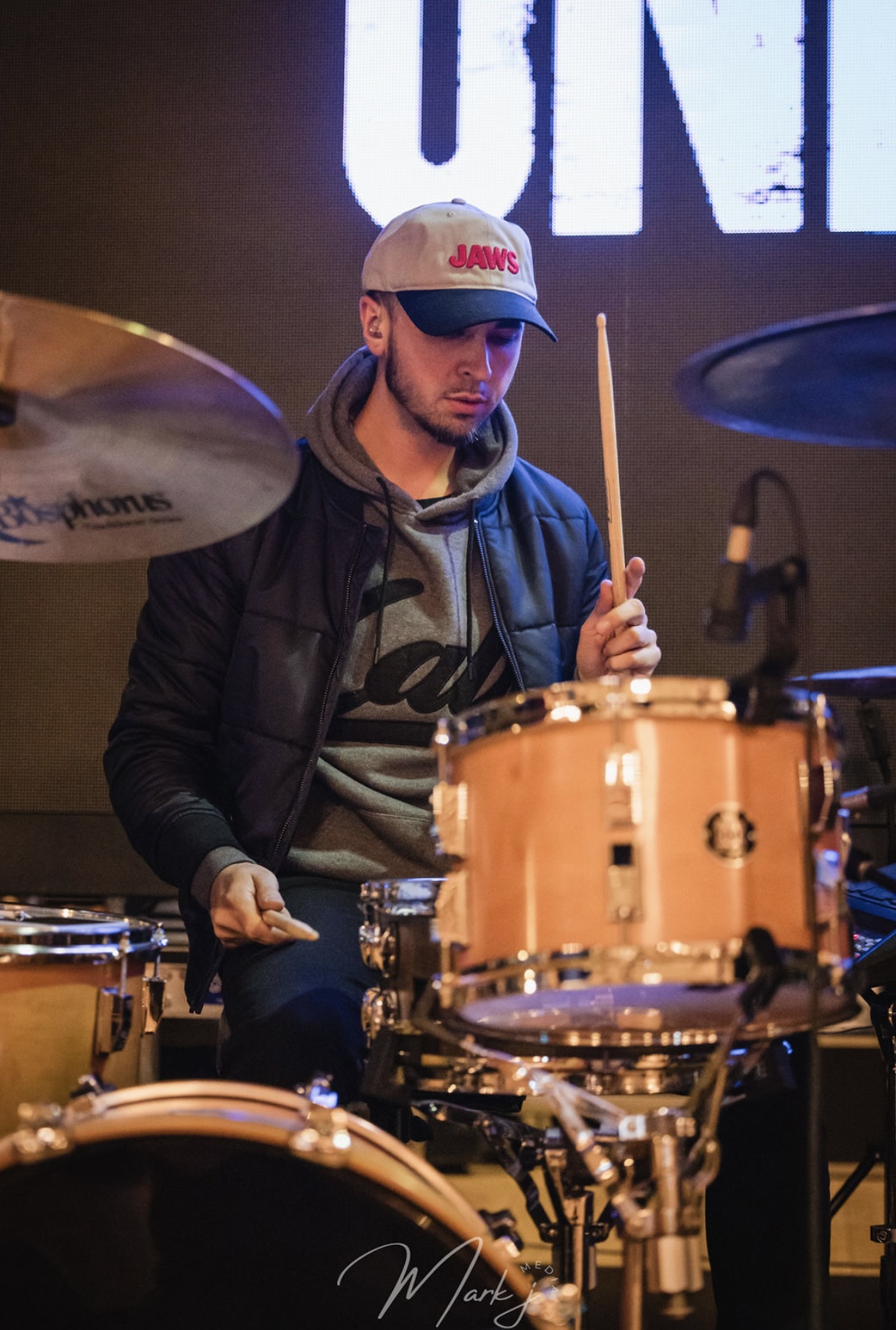 Young man playing drums on stage, wearing a cap and hoodie, with a large screen behind him.