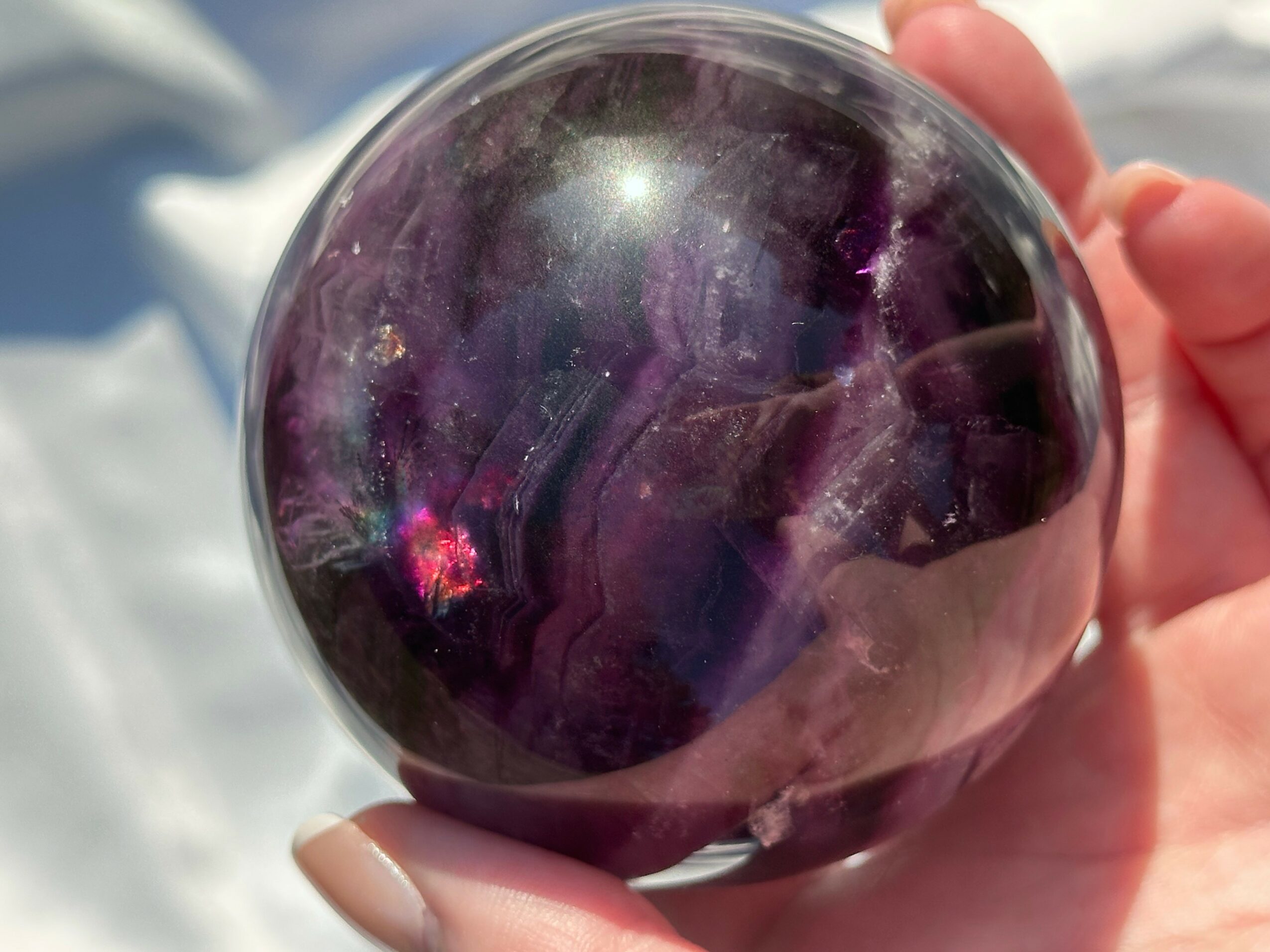 Hand holding a shiny, dark purple sphere with reflections and light spots, outdoors with bright background.