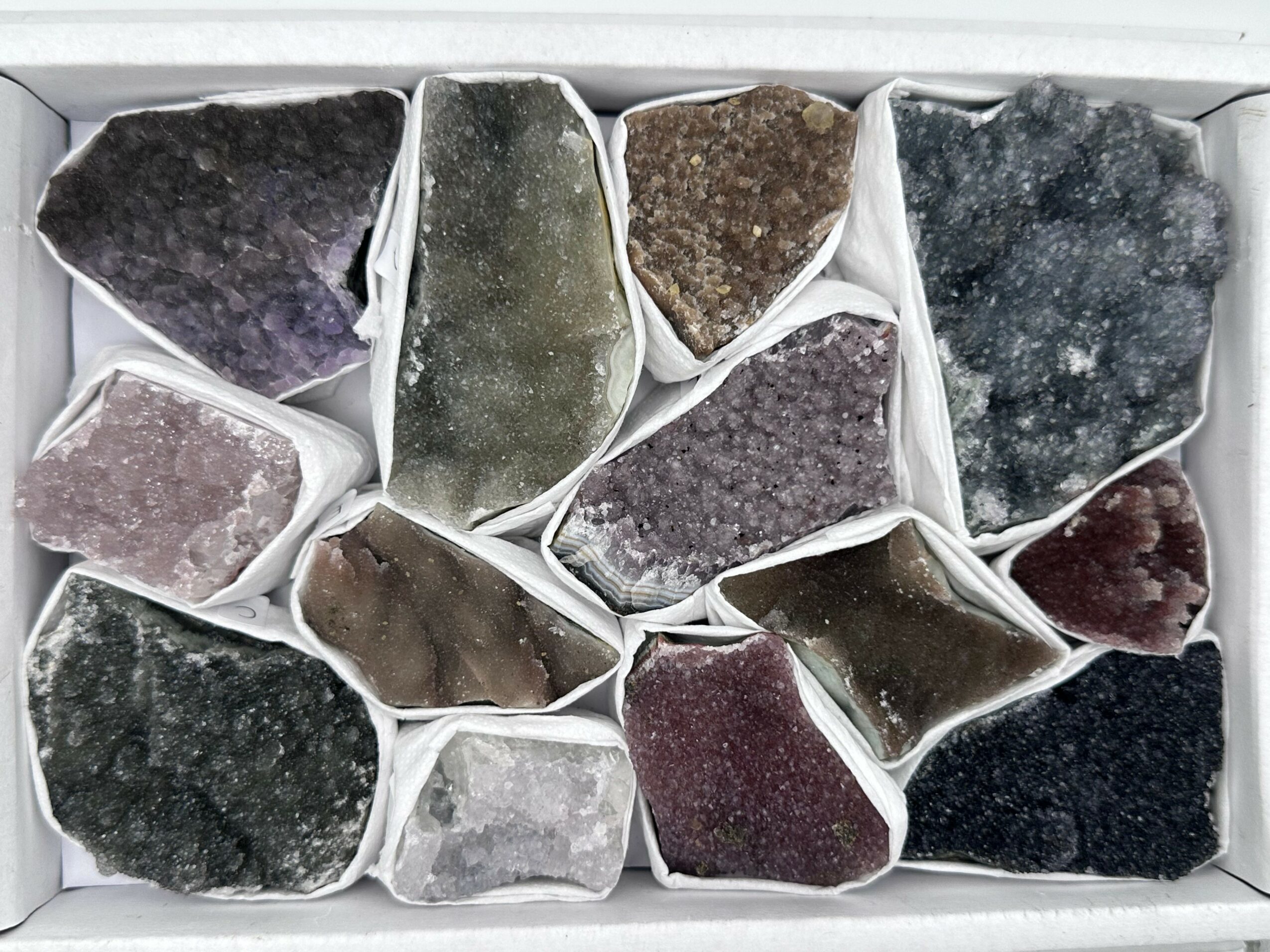 Collection of various colored mineral or gemstone specimens in small white containers, arranged in a box.