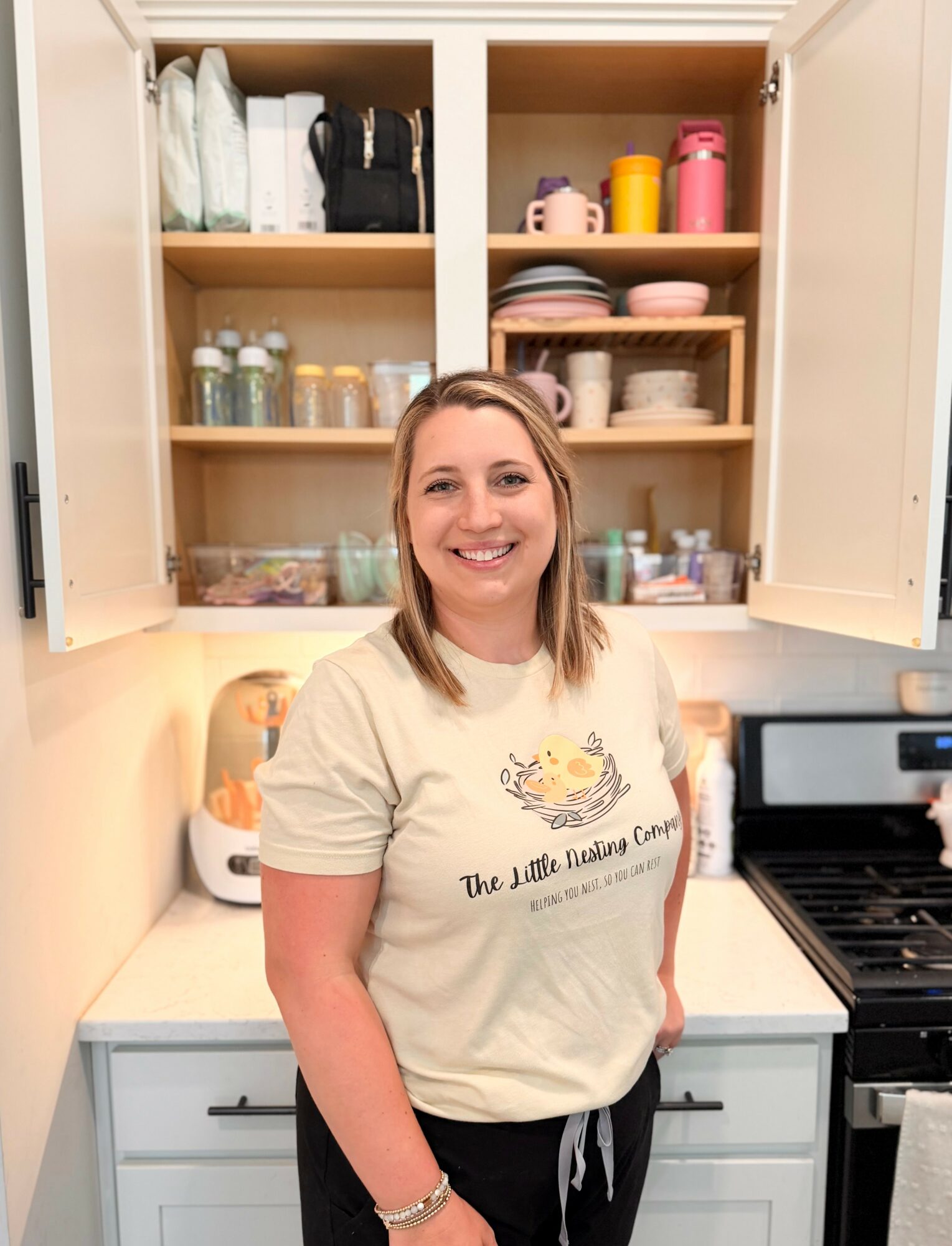 Smiling woman standing in a kitchen with open cabinets filled with dishes and containers, wearing a light-colored T-shirt.