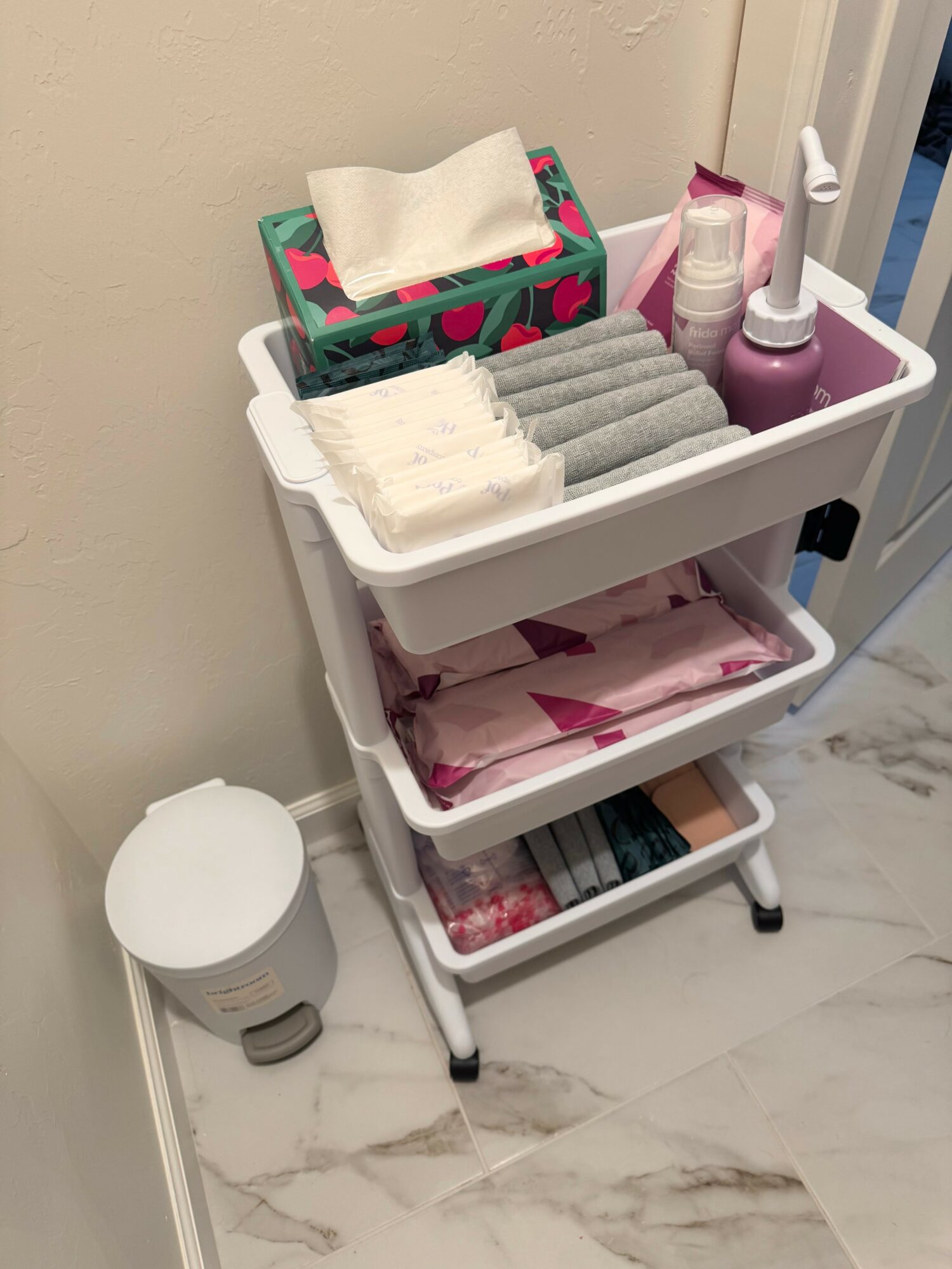Three-tier white cart with towels, bottles, and a tissue box, next to a small white trash can on marble floor.