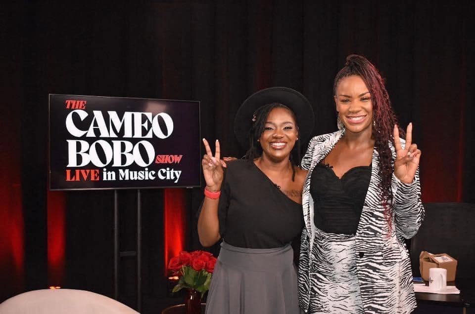 Two women smiling and making peace signs, standing in front of a sign that reads 'The Cameo Bobo Show Live in Music City.'