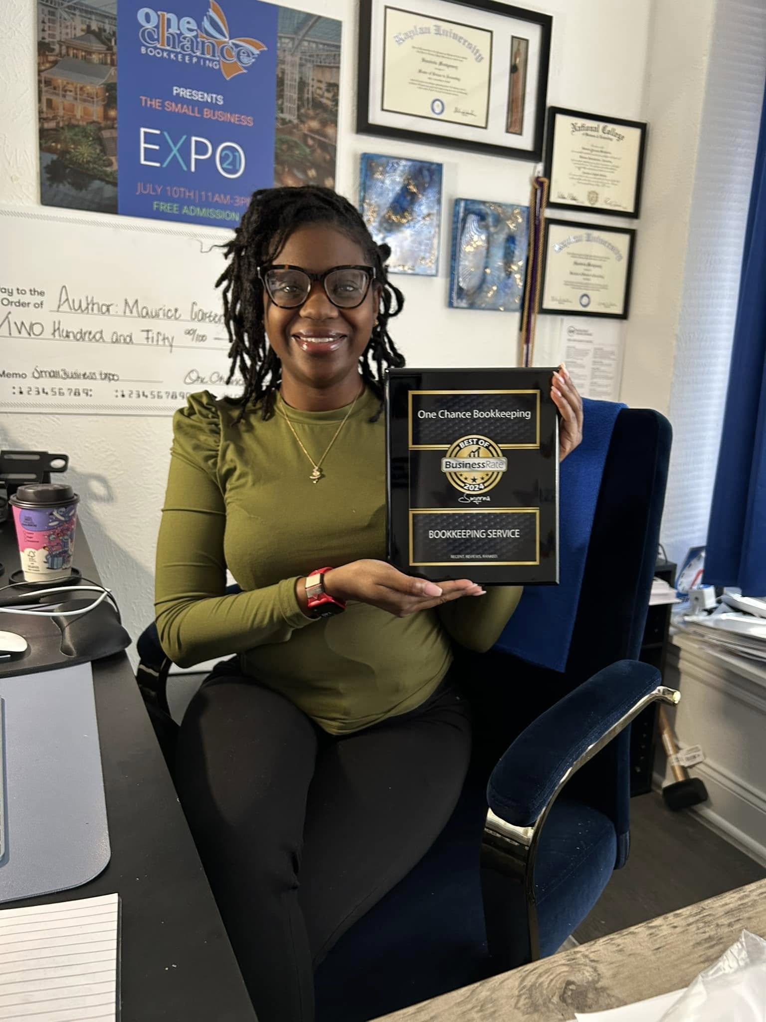 Woman with glasses and dreadlocks smiling, holding a plaque in an office with certificates and awards on the wall.