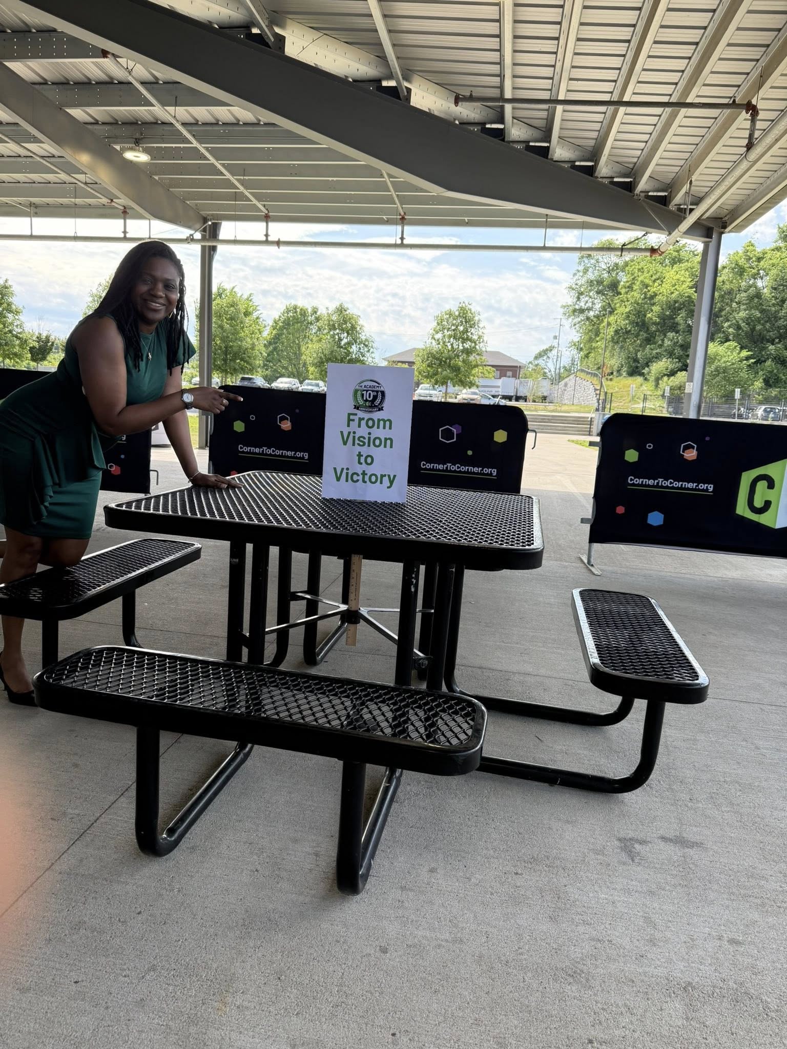 Girl standing at picnic table with computers, outdoors with trees and sky in background.