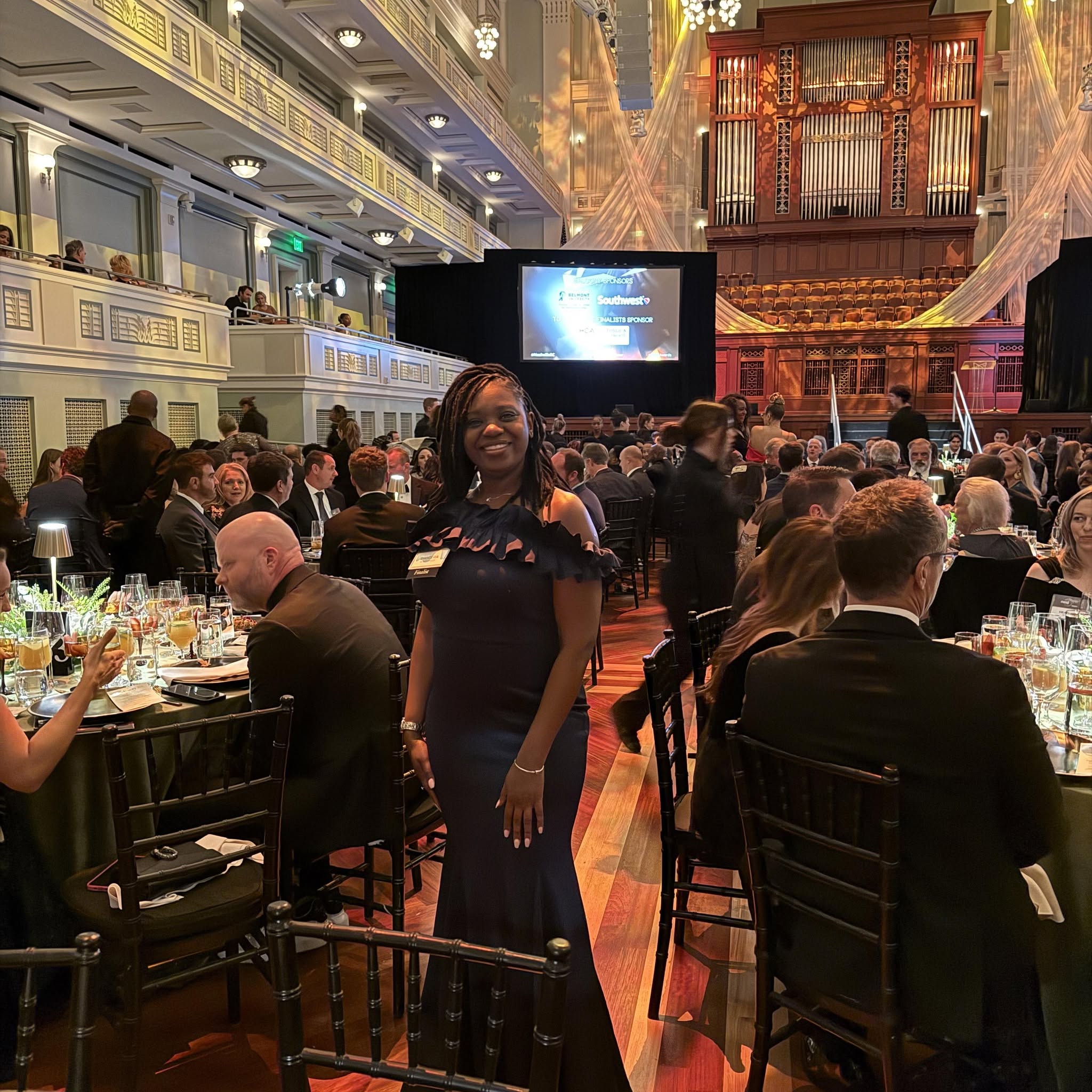 Woman in a dark dress standing in a large banquet hall filled with seated guests, tables, and a large screen on stage.