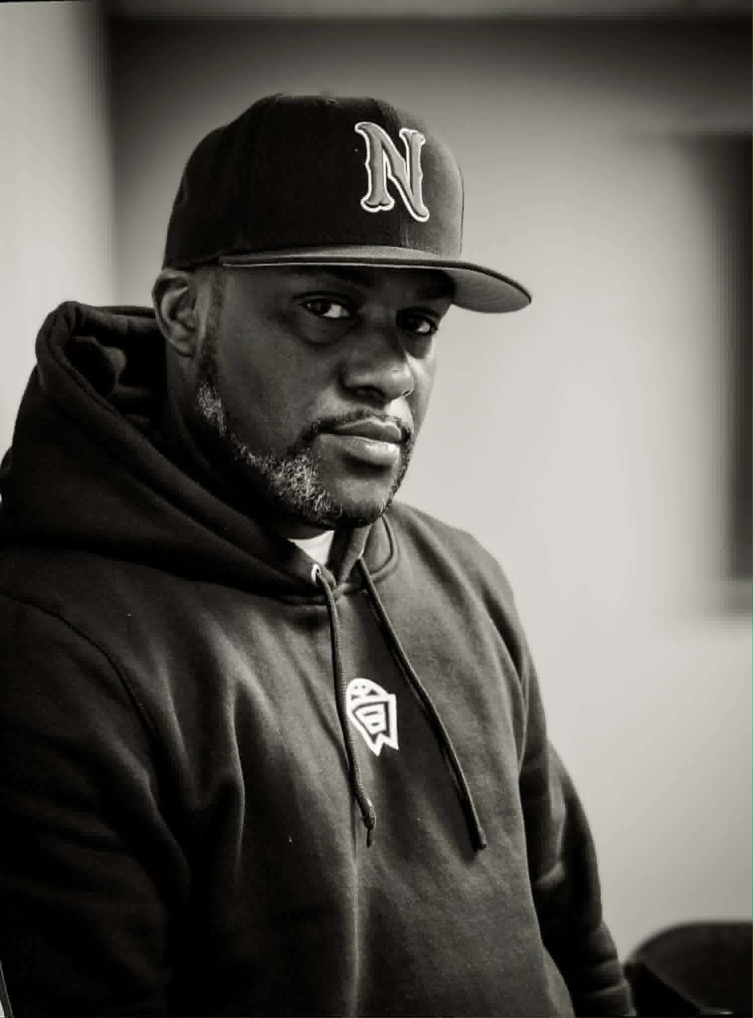 Man wearing a baseball cap with an 'N' logo and a hoodie, looking at the camera in black and white.