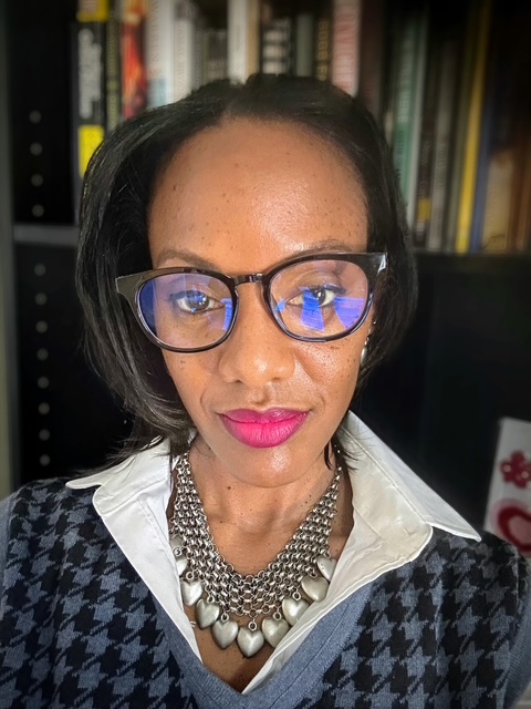 Close-up of a woman with glasses, wearing a patterned sweater and necklace, in front of a bookshelf.