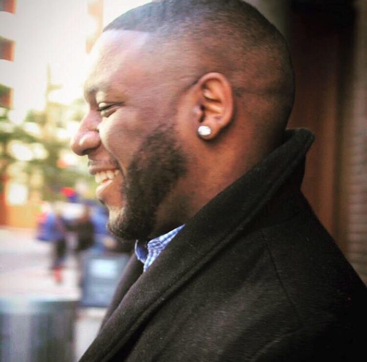 Side profile of smiling man with short hair, beard, and earring, outdoors in an urban setting.