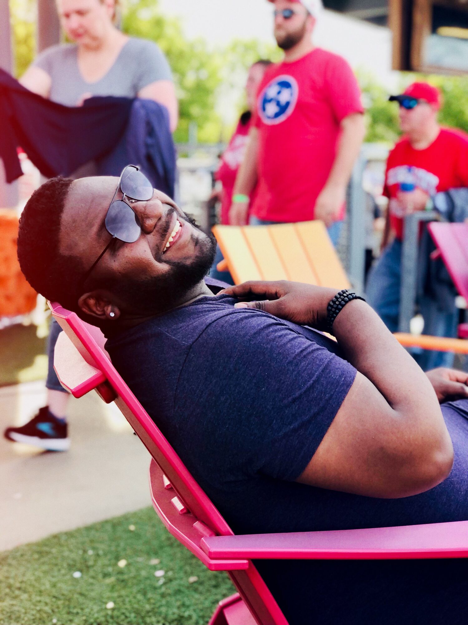 Person with sunglasses relaxing on a pink lounge chair outdoors, smiling with arms crossed, surrounded by people in casual clothing.