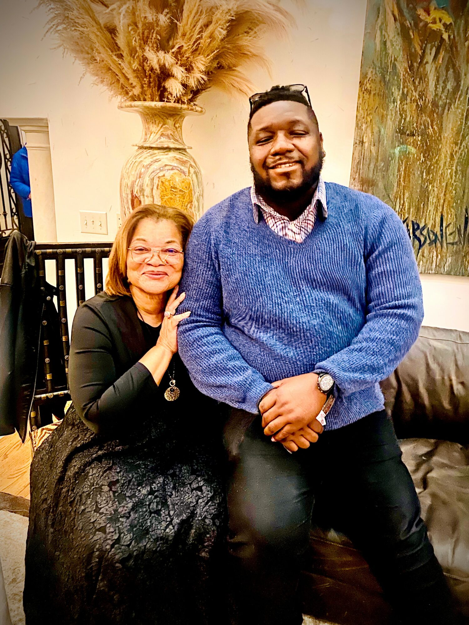 A woman and a man sit together indoors, smiling at the camera, with decorative art and a large vase in the background.