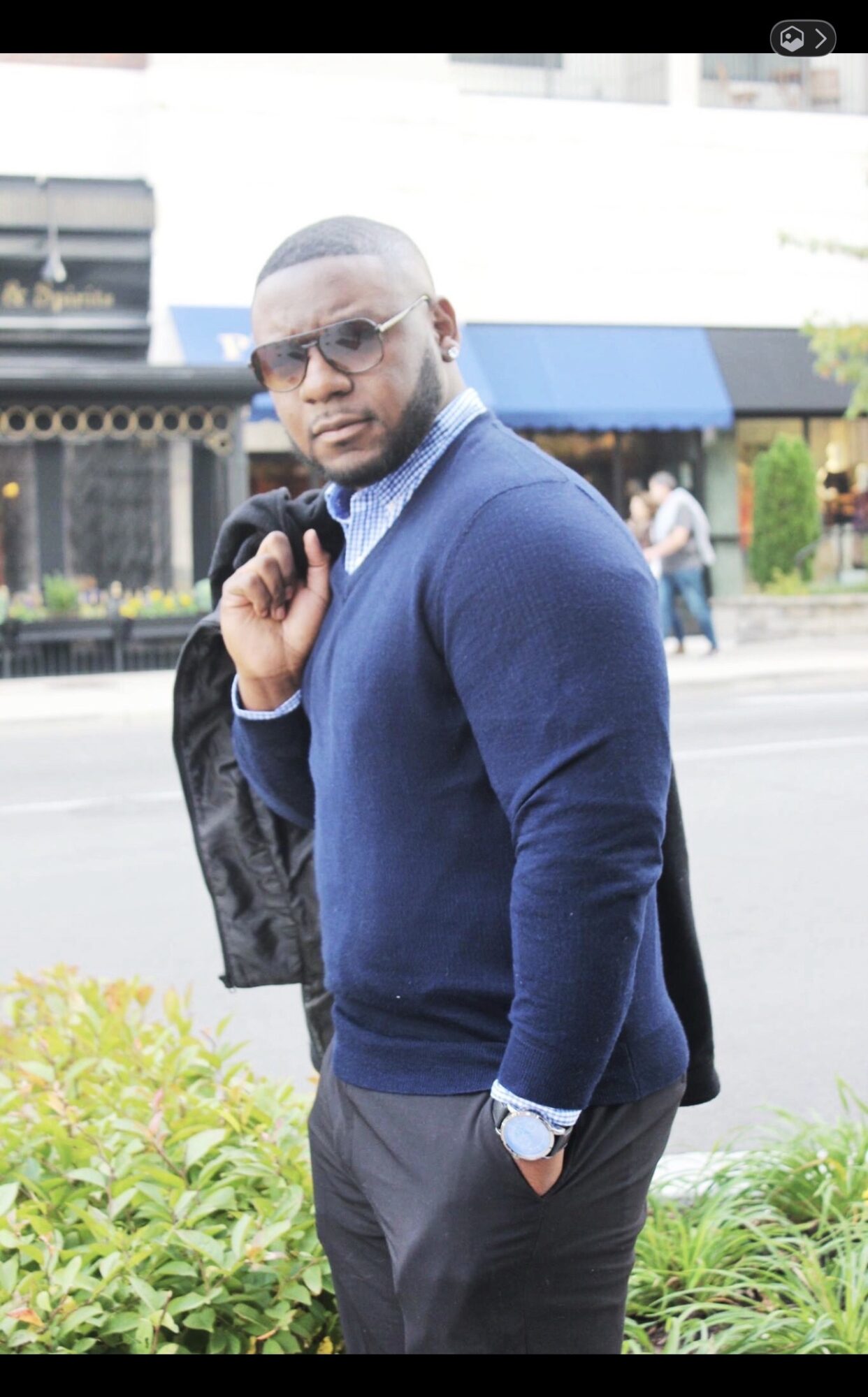 Man in blue sweater and sunglasses standing outdoors with a jacket over his shoulder, hands in pockets, in front of a street scene