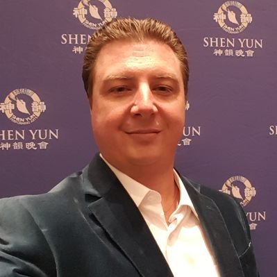 Man in suit smiling in front of a purple backdrop with Chinese characters and the words 'SHEN YUN'.