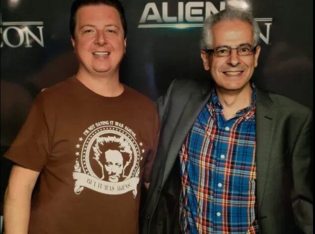 Two men smiling, standing in front of a dark background with the word 'ALIEN' visible, one wearing a brown T-shirt and the other a plaid shirt and blazer.