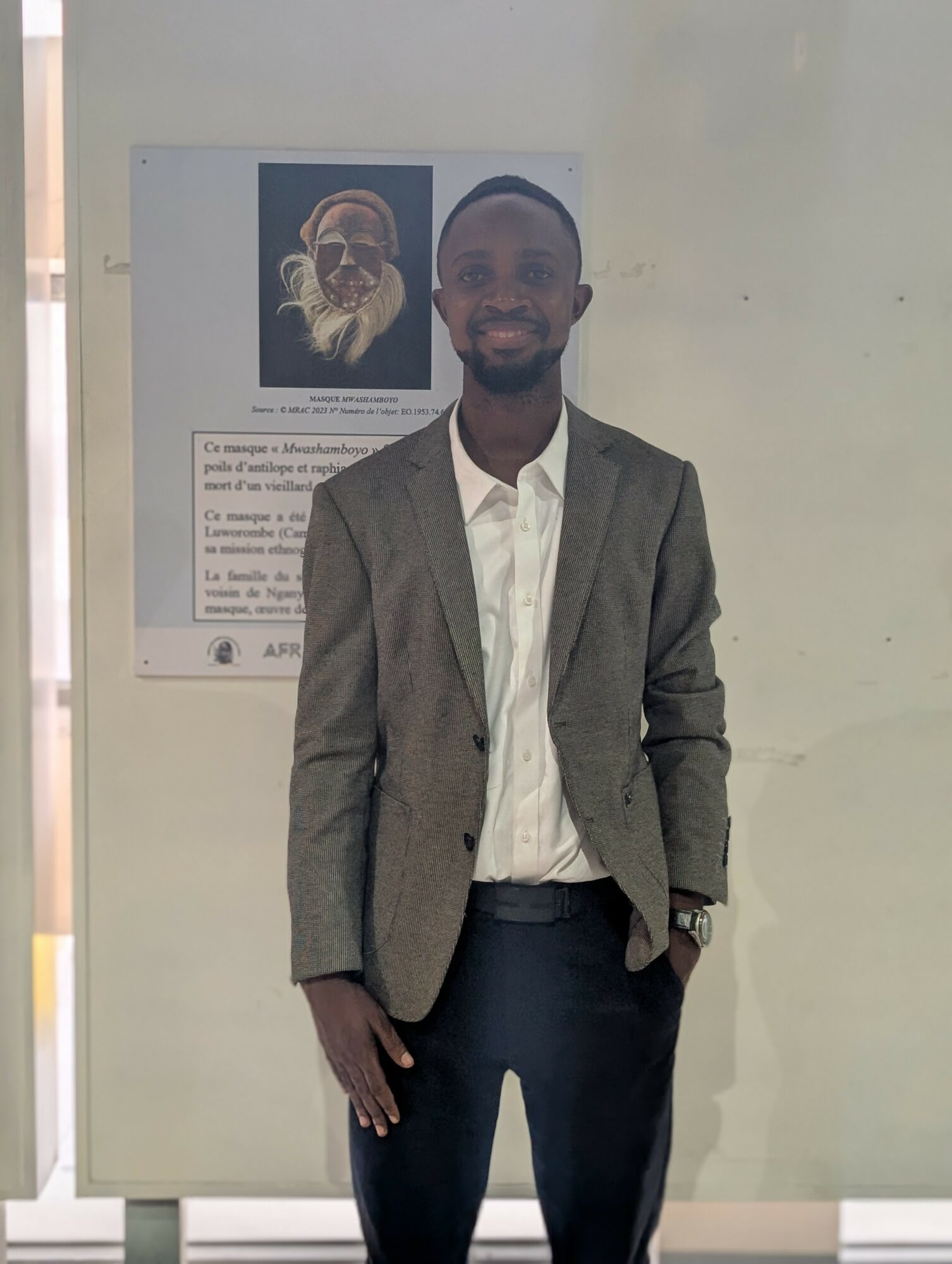 Young man in a gray blazer and white shirt standing indoors, smiling, with a poster on the wall behind him.