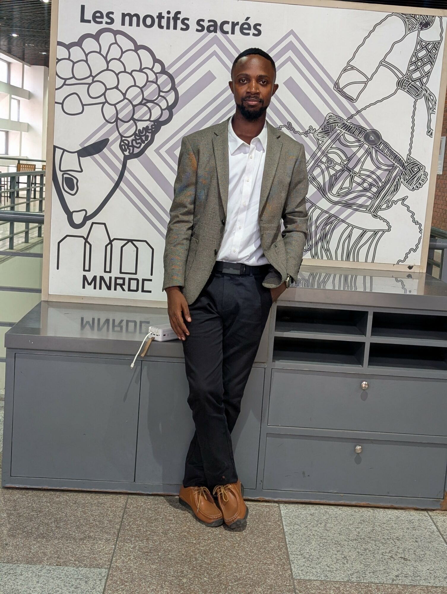 Man standing in front of a mural with abstract drawings and text, wearing a blazer, white shirt, and dark pants.