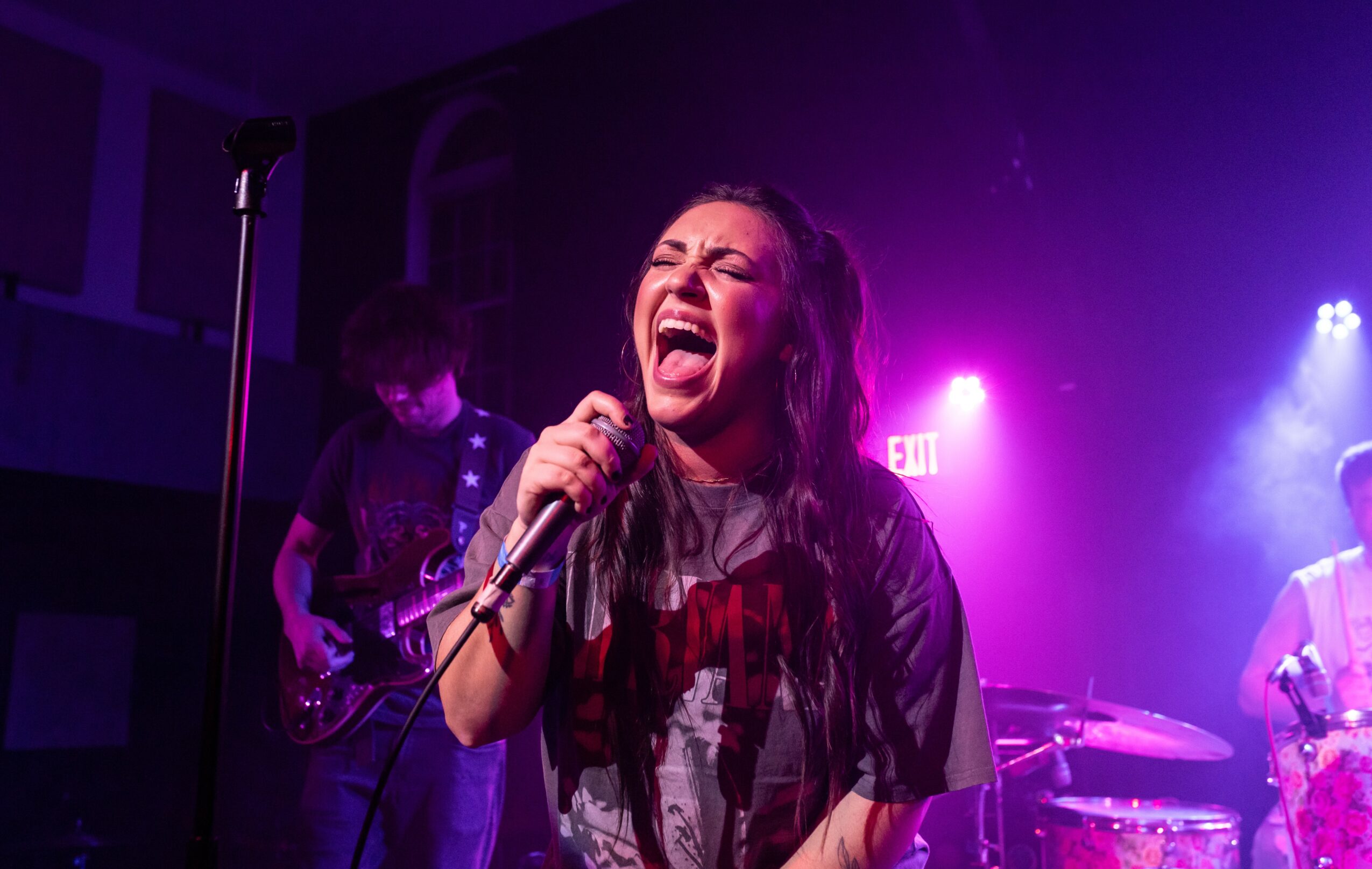 Female singer passionately performs on stage with band, purple and blue lighting, holding microphone, expressive face.