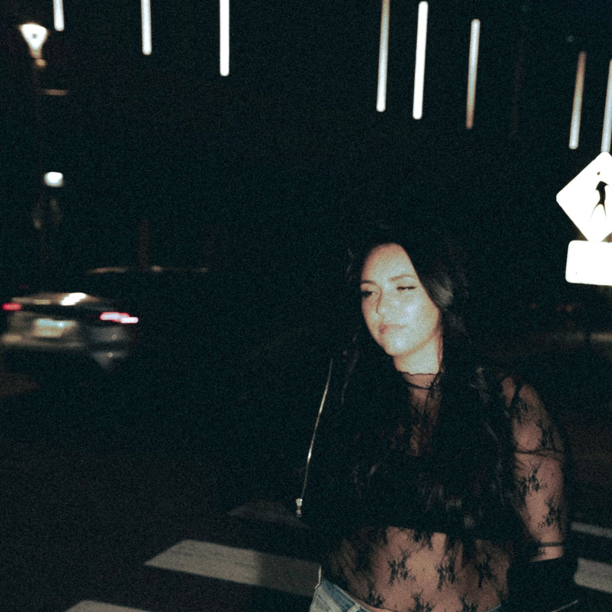 Woman with long dark hair standing outdoors at night, illuminated by streetlights, with a dark background and blurred lights.