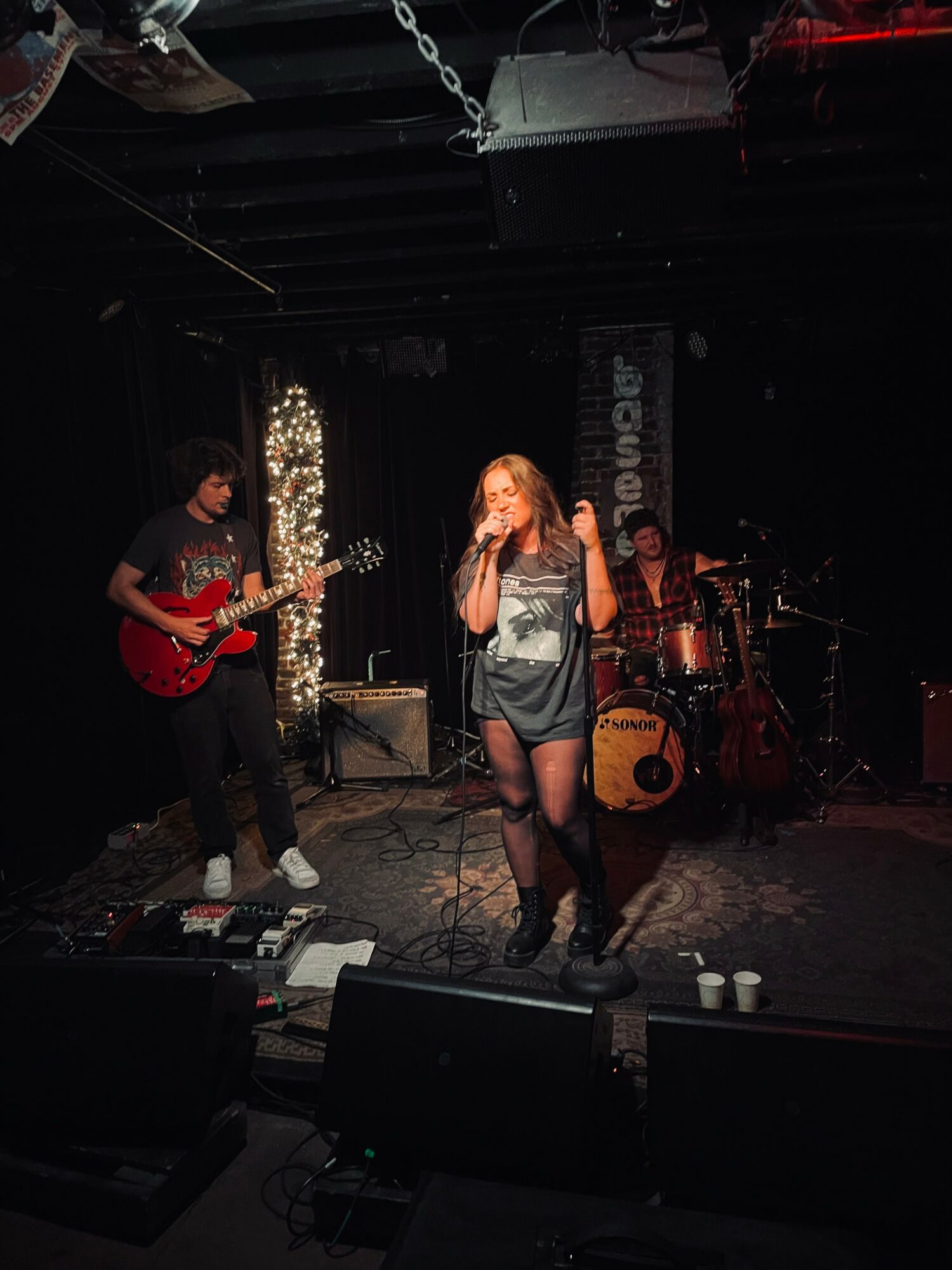 Three musicians perform on stage with a female singer in the center, a guitarist on the left, and a drummer on the right.
