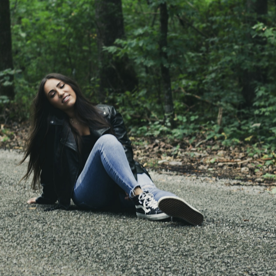 Young woman with long dark hair sitting on a paved path in a forest, wearing a black leather jacket, jeans, and sneakers.