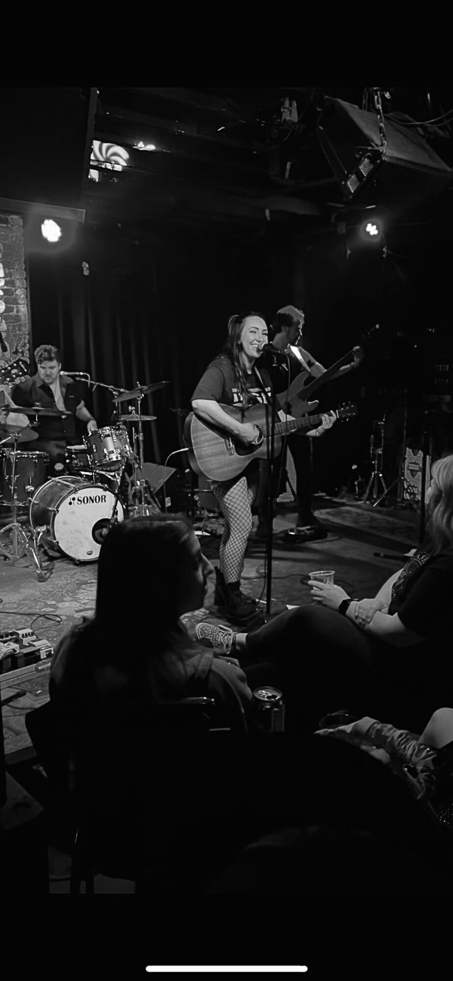 Band performs live on stage with a drummer, guitarist, and bassist, audience seated in front, dim lighting, intimate setting.