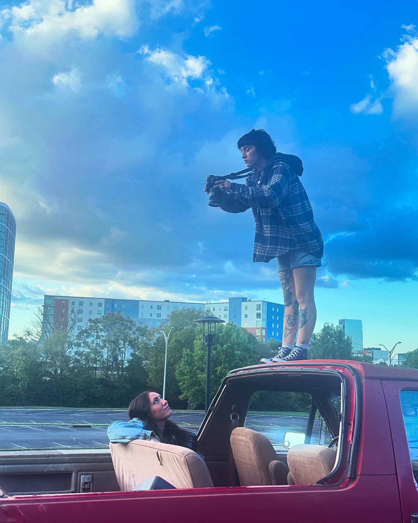 Person standing on a car's roof taking a photo of a woman sitting inside the vehicle, with a cityscape and cloudy sky in background.