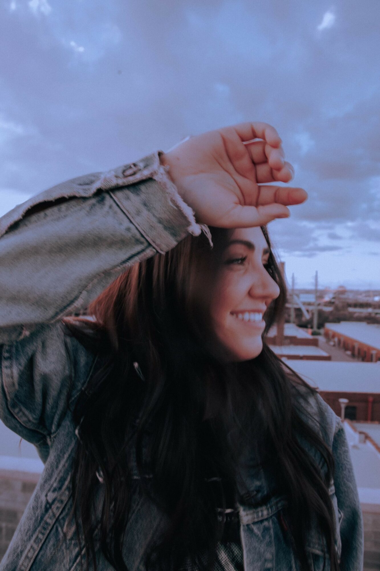 Young woman smiling outdoors with her hand raised above her forehead, sky and buildings in background.