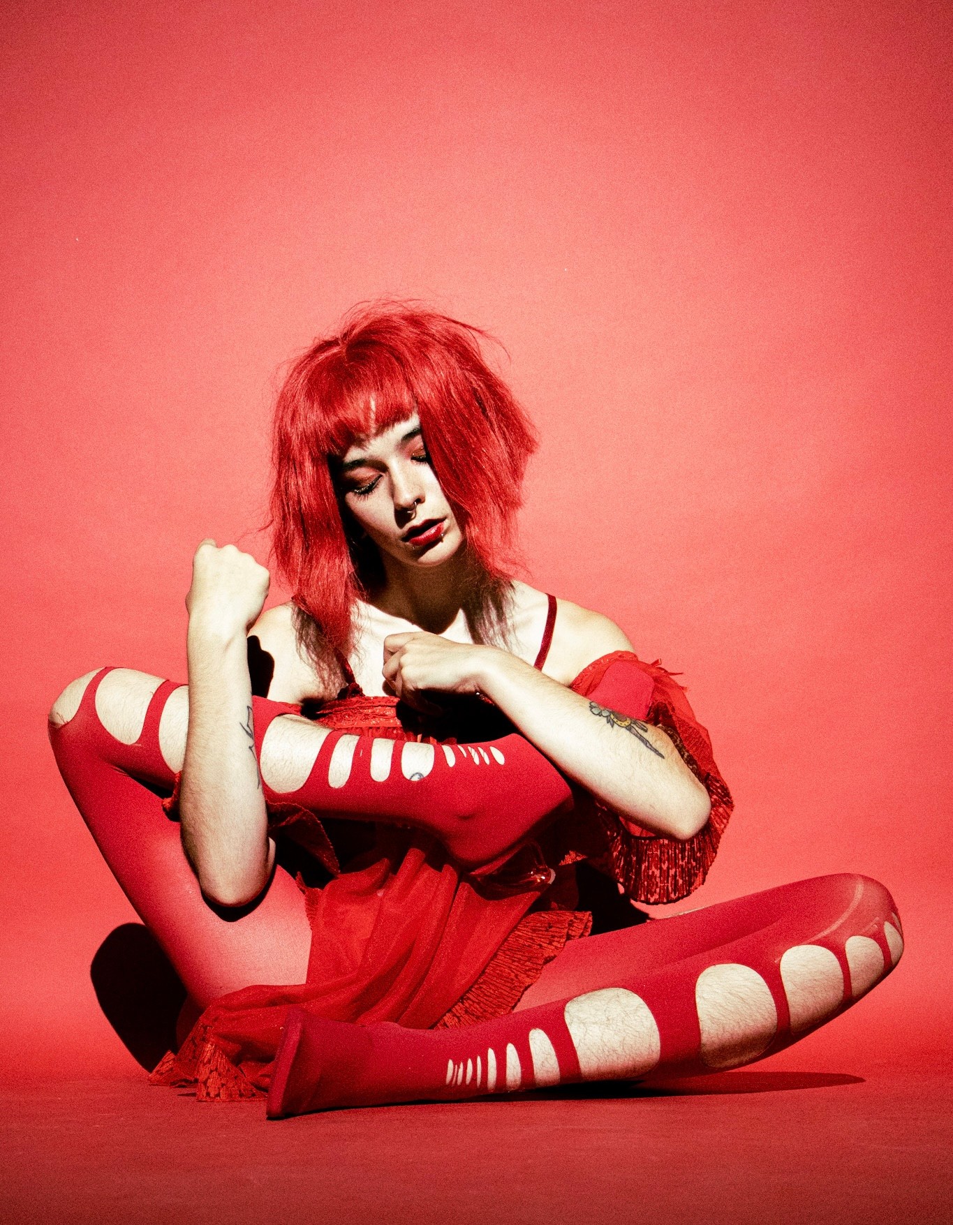 Person with red hair and makeup sitting on the floor in a red outfit against a red background.
