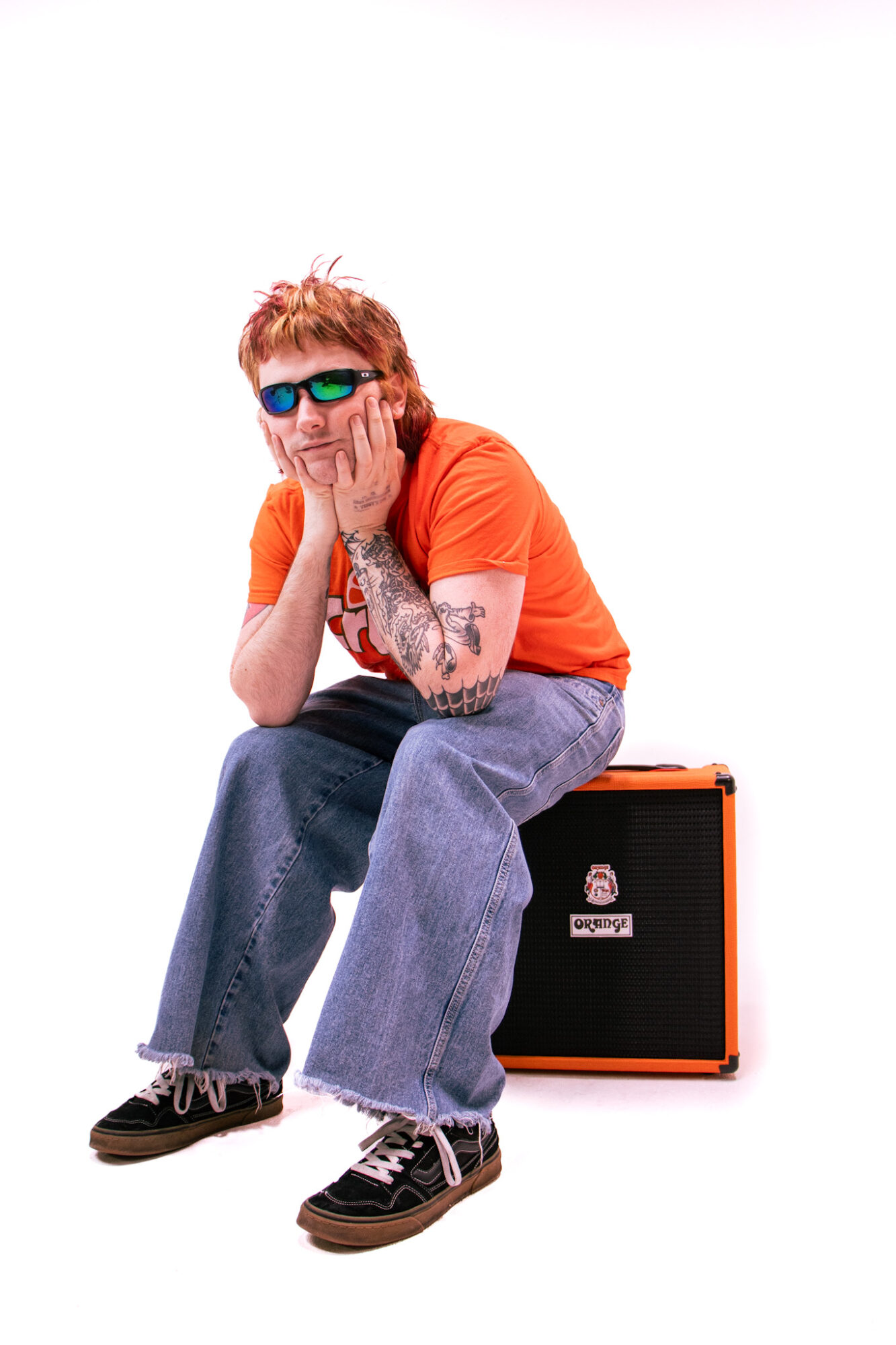 Person with red hair, sunglasses, orange shirt, and tattoos sitting on an orange amplifier, smiling with hands on face.