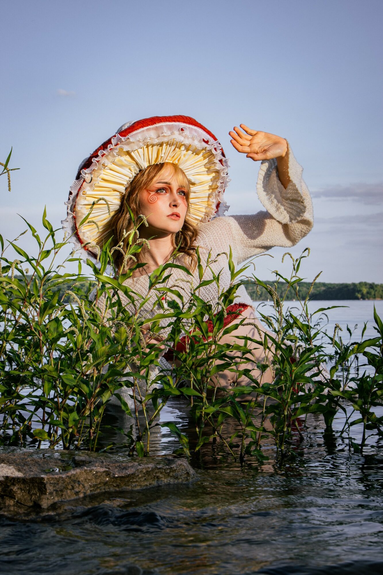 Young woman wearing a large, colorful hat and light clothing, standing among plants near water, raising her hand.