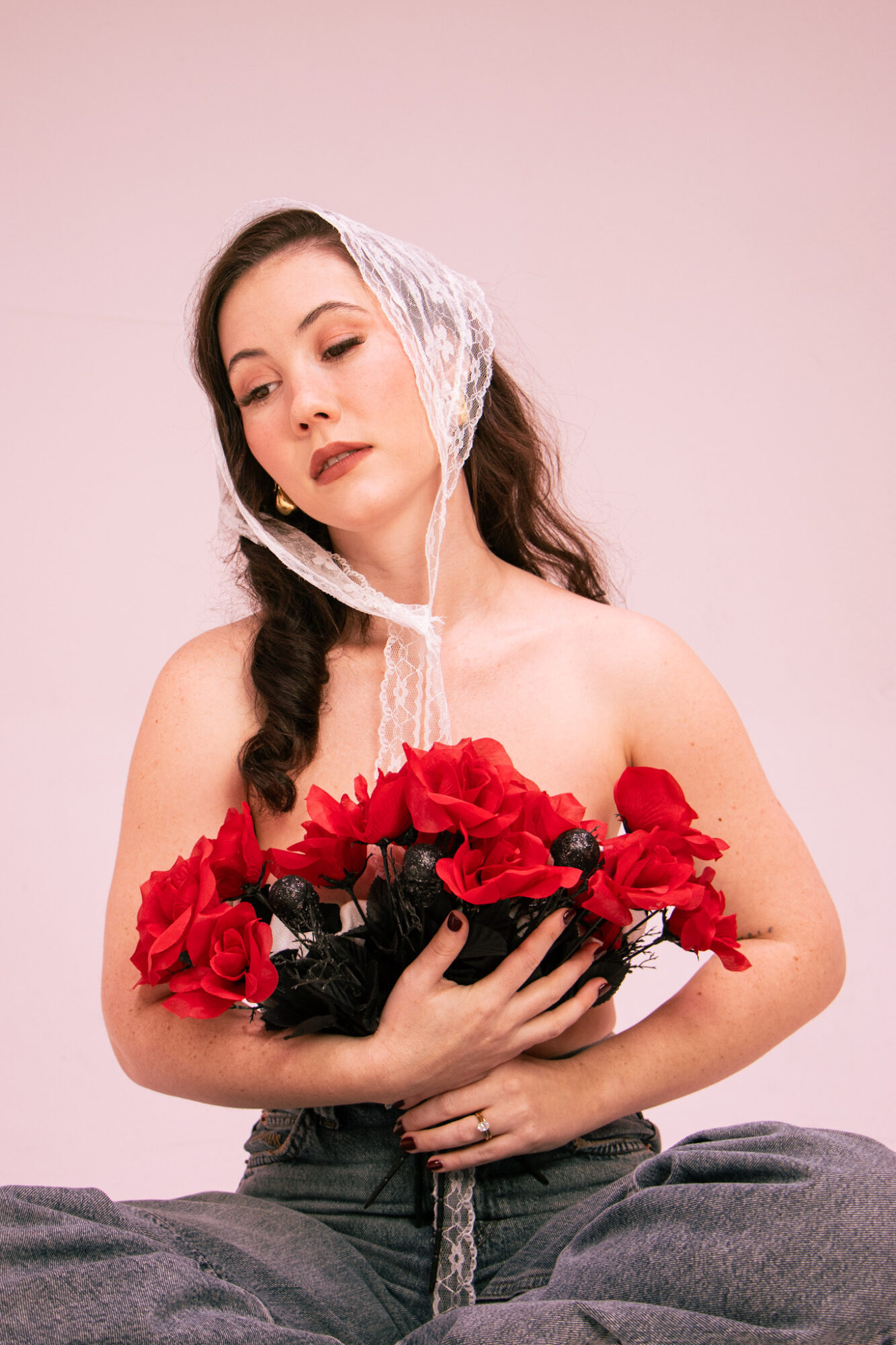 Young woman with dark hair, wearing a lace headscarf, holding red flowers, sitting against a pink background.
