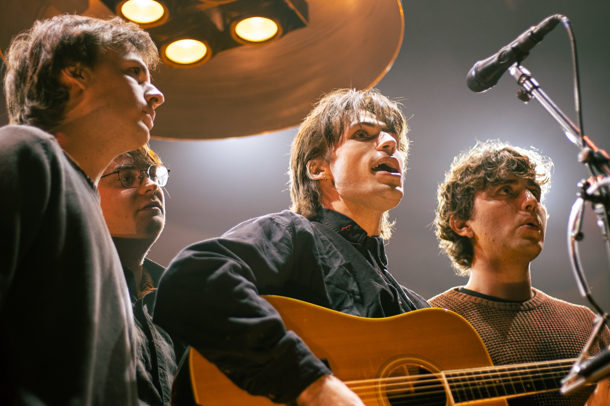 Four people singing and playing guitar on stage with a microphone, illuminated by stage lights.