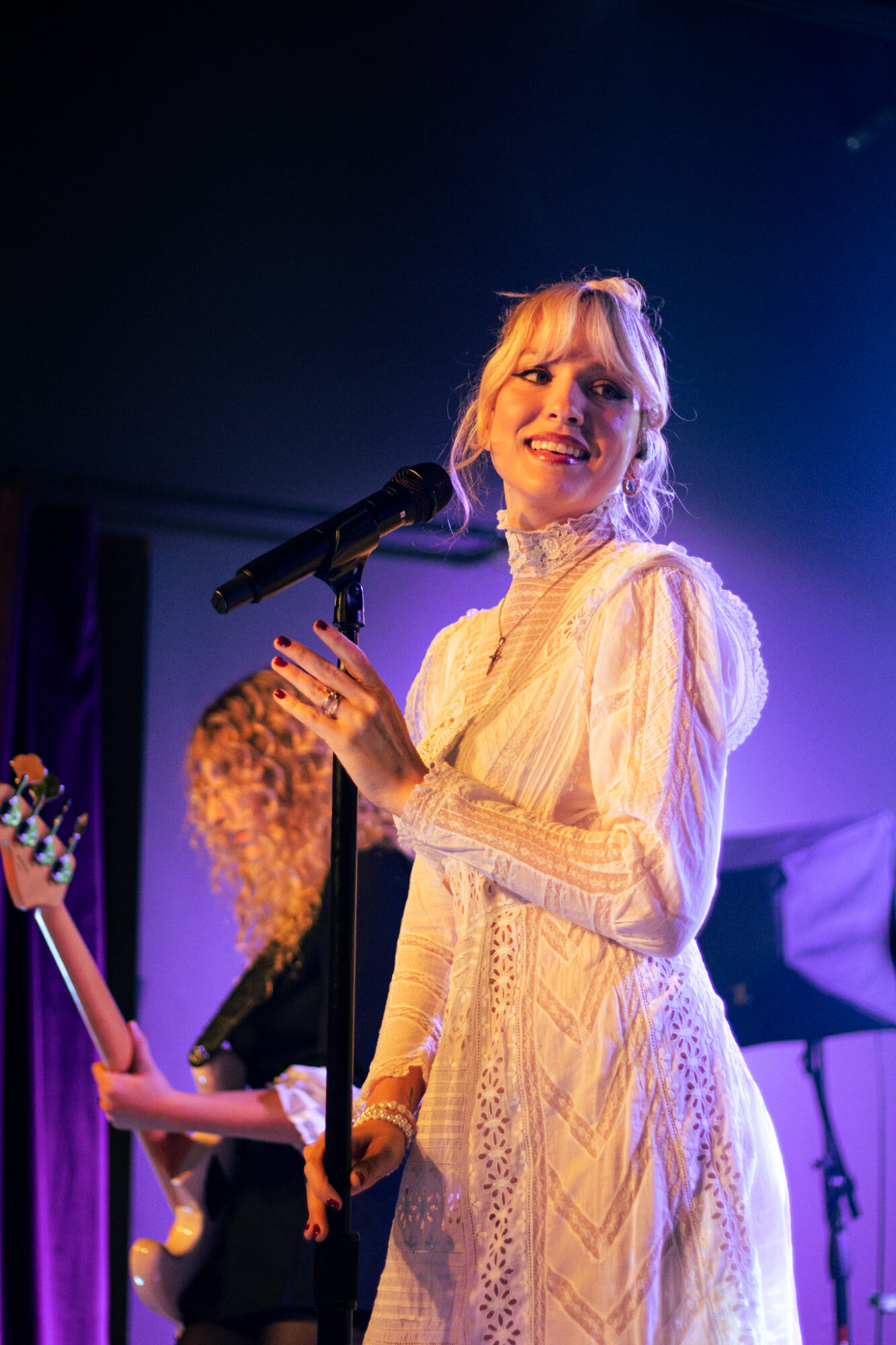 Woman singing into a microphone on stage, wearing a light-colored dress, with a band in the background.