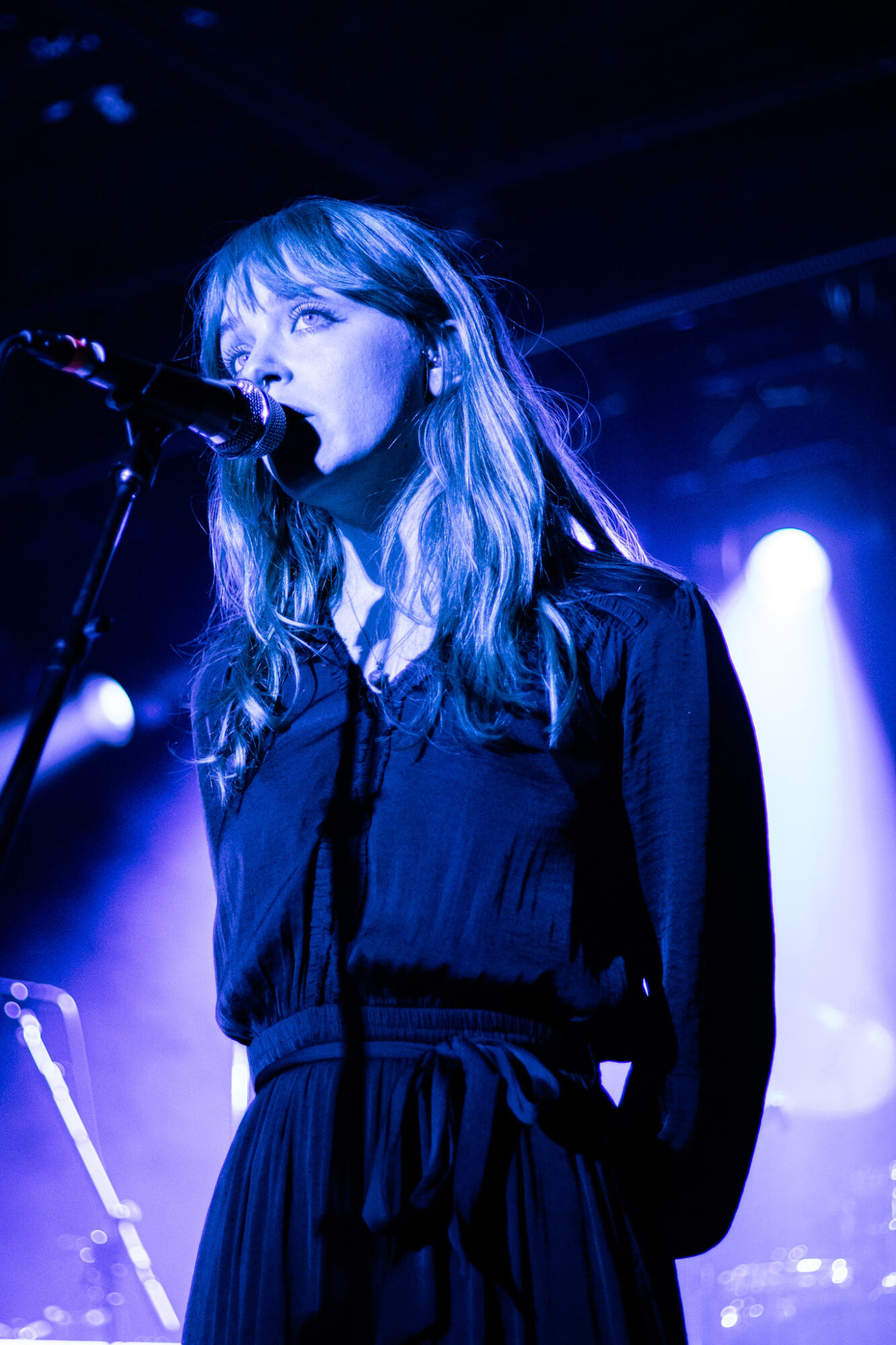 Female singer with long hair performs on stage with blue lighting, singing into a microphone.