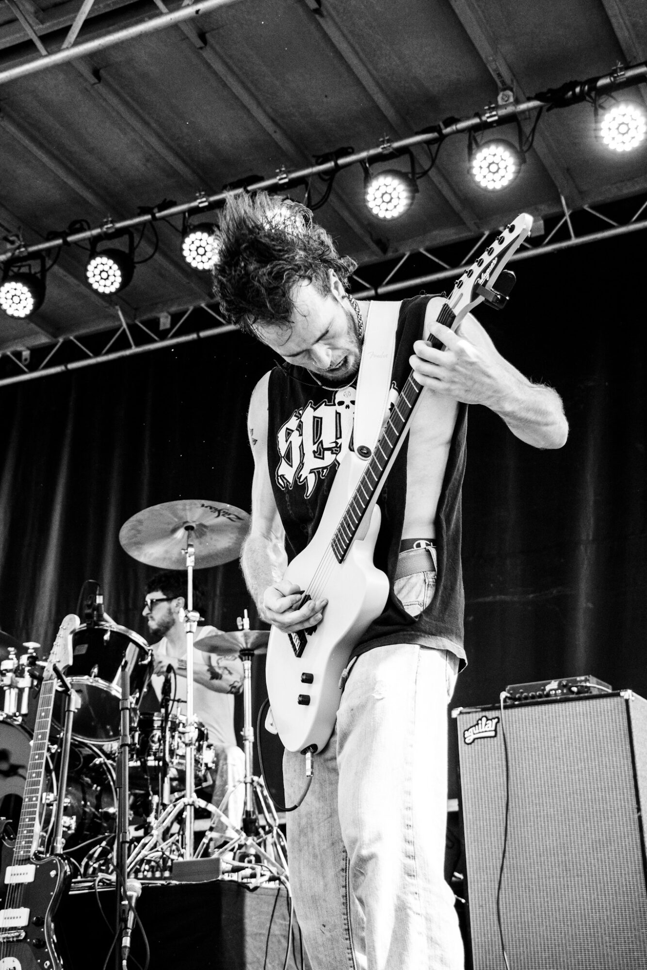 Musician playing an electric guitar on stage with drums and stage lights in background.