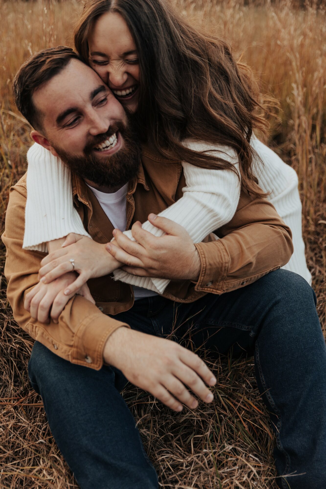 A smiling couple hugging outdoors in a field with tall grass, the woman on the man's back, both laughing.